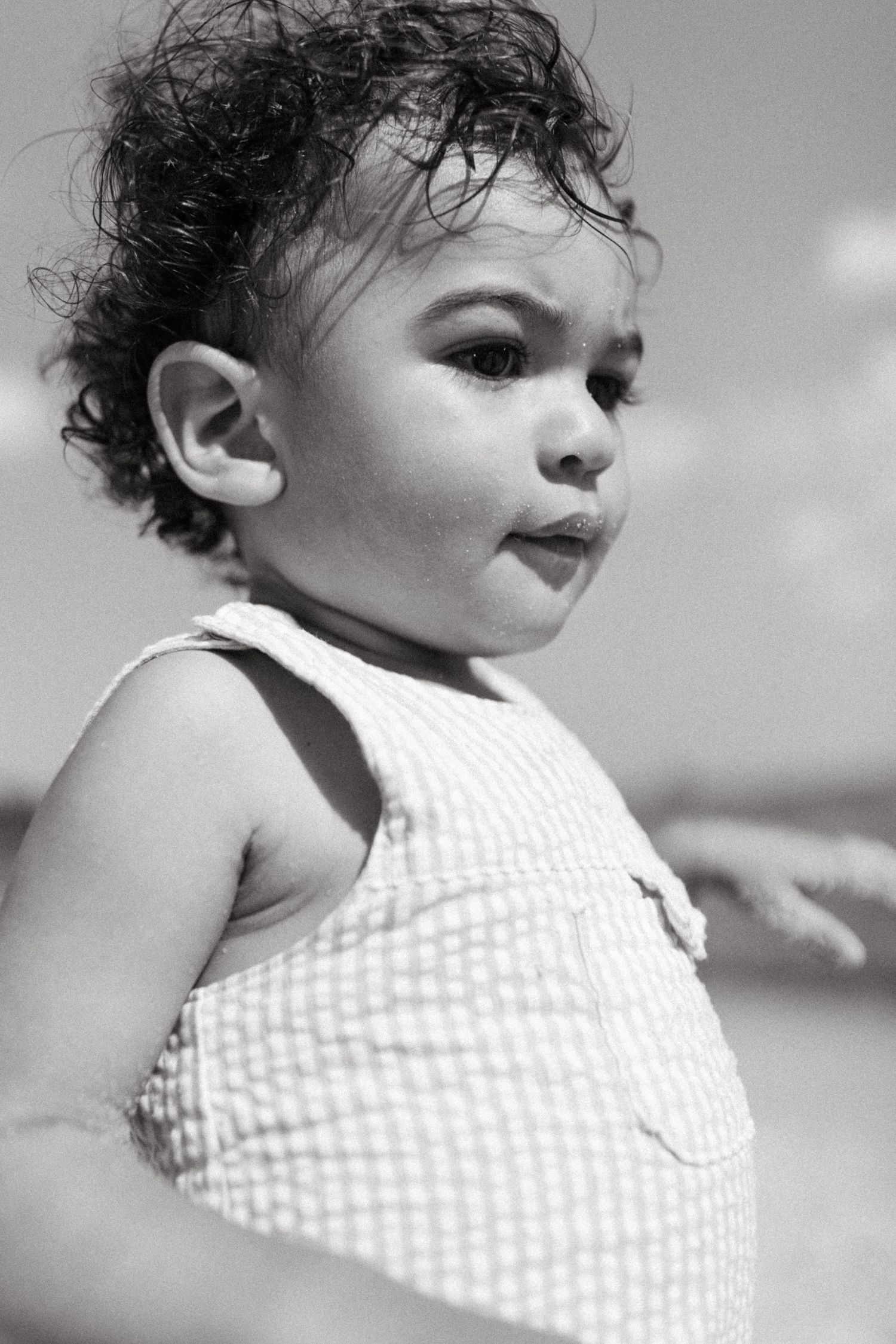Black and white portrait of a child with curly hair and a checkered sundress looking to the side contemplatively.
