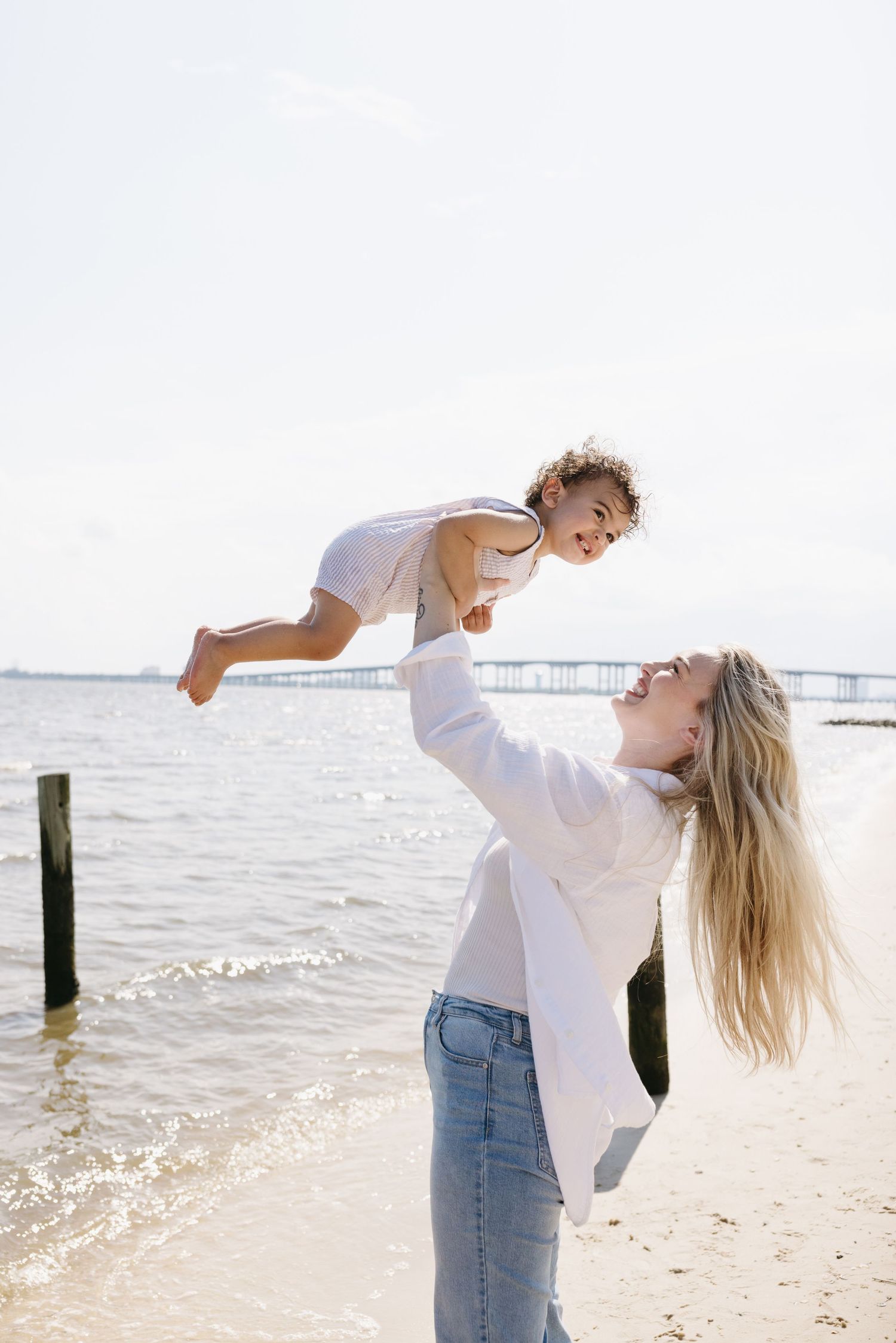 A parent holds and lifts a small child playfully at the beach with ocean waves and pier visible in background.