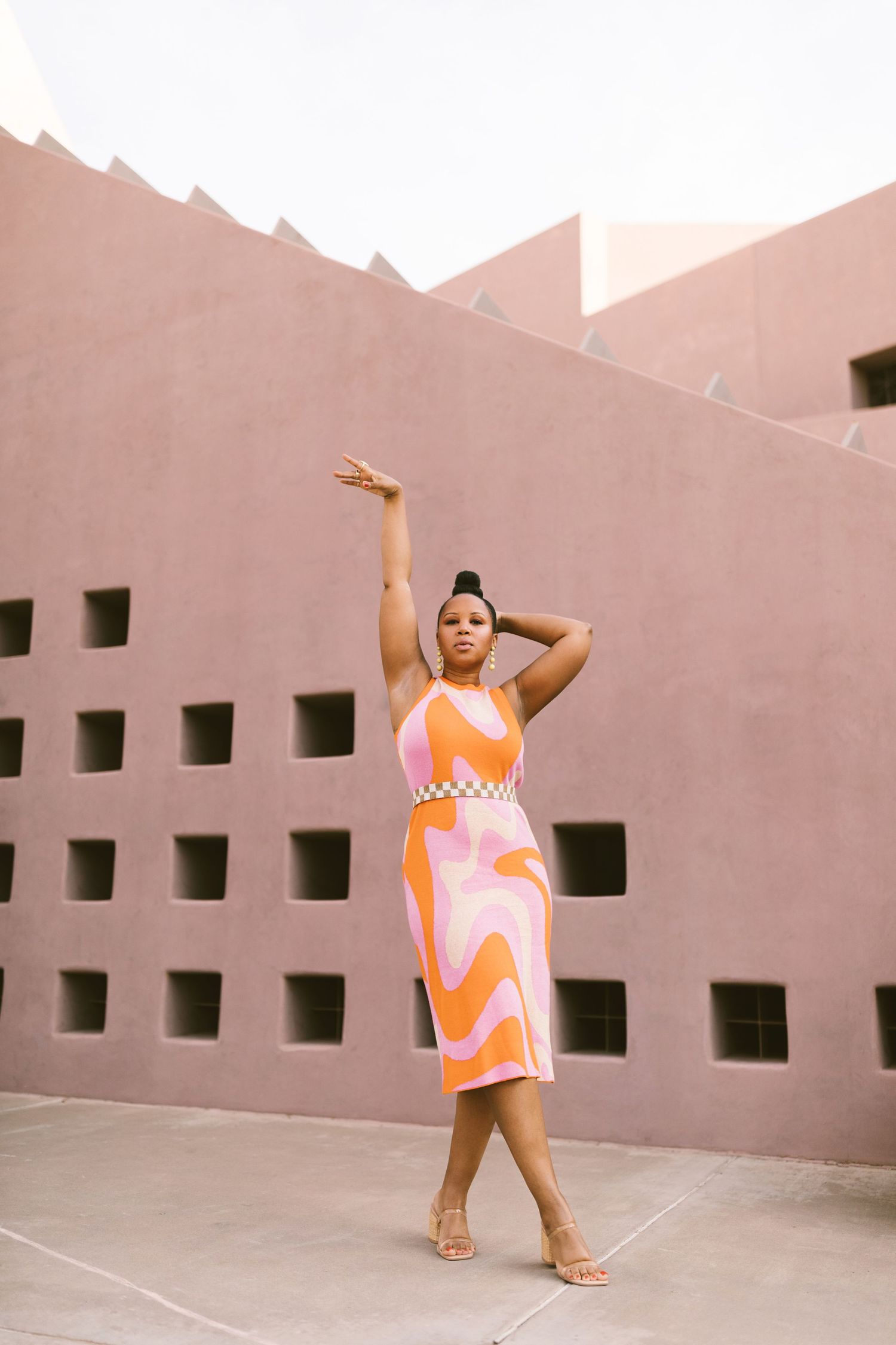 A person poses gracefully against a pink wall wearing a vibrant orange and white patterned sleeveless dress and sandals.