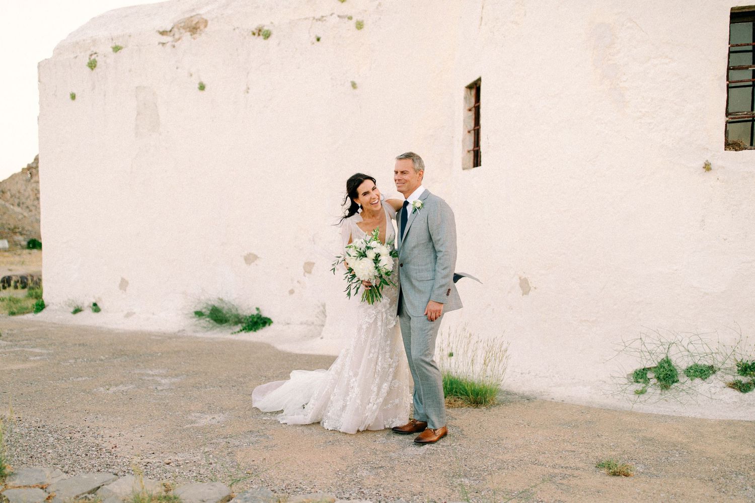 A couple in wedding attire pose together against a white adobe wall in a desert landscape.