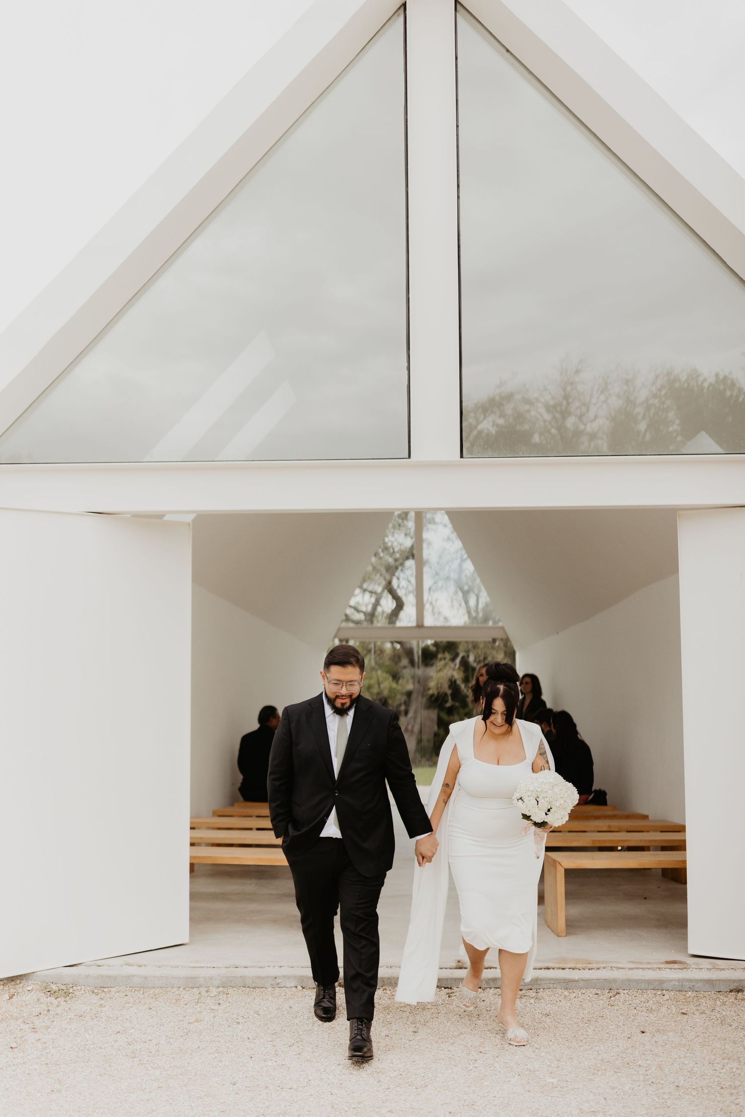 A wedding couple walks hand in hand exiting a modern white chapel with triangular windows.