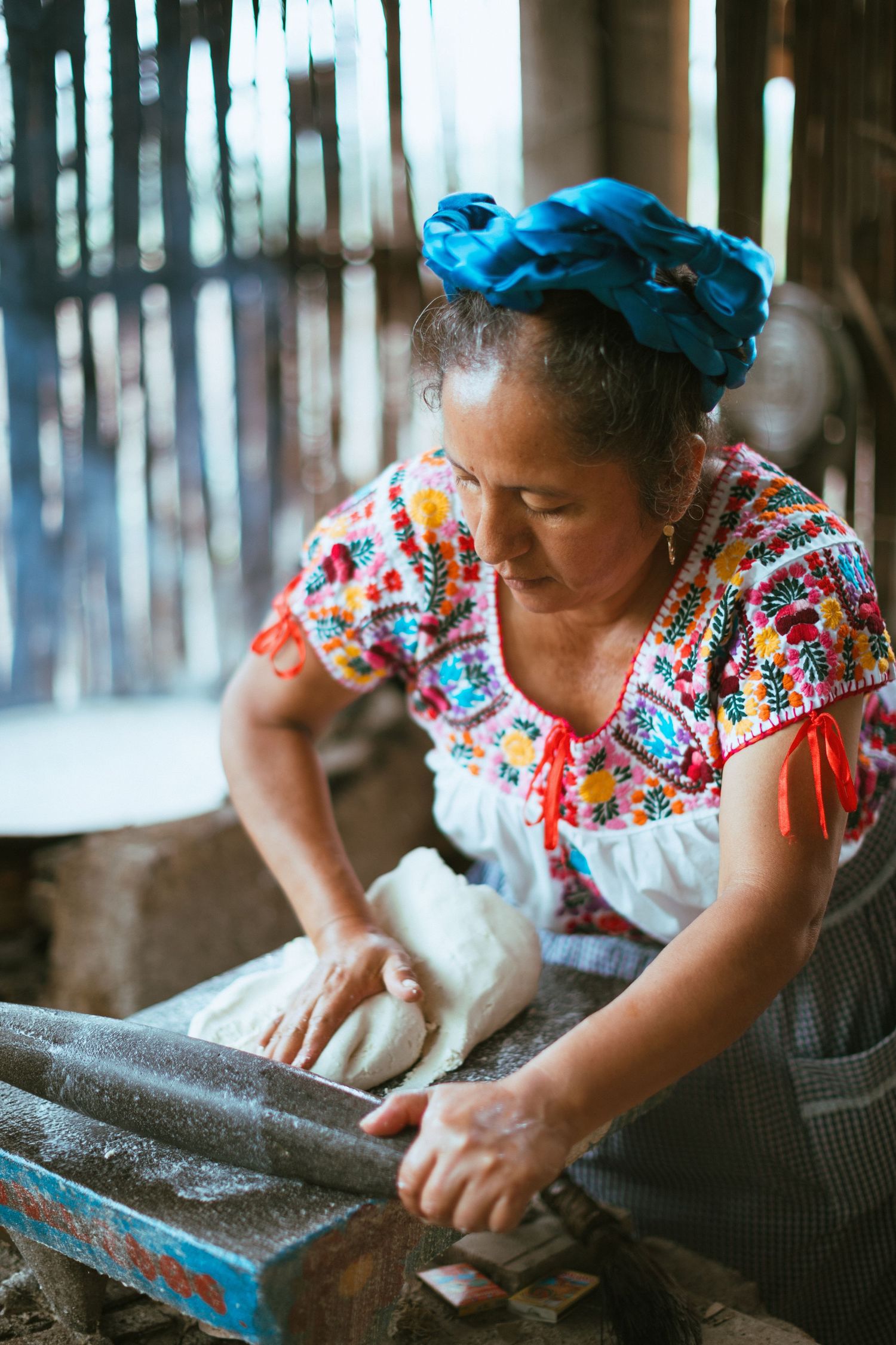 A person wearing a colorful embroidered blouse kneads dough on a rustic wooden surface while working in a traditional kitchen.
