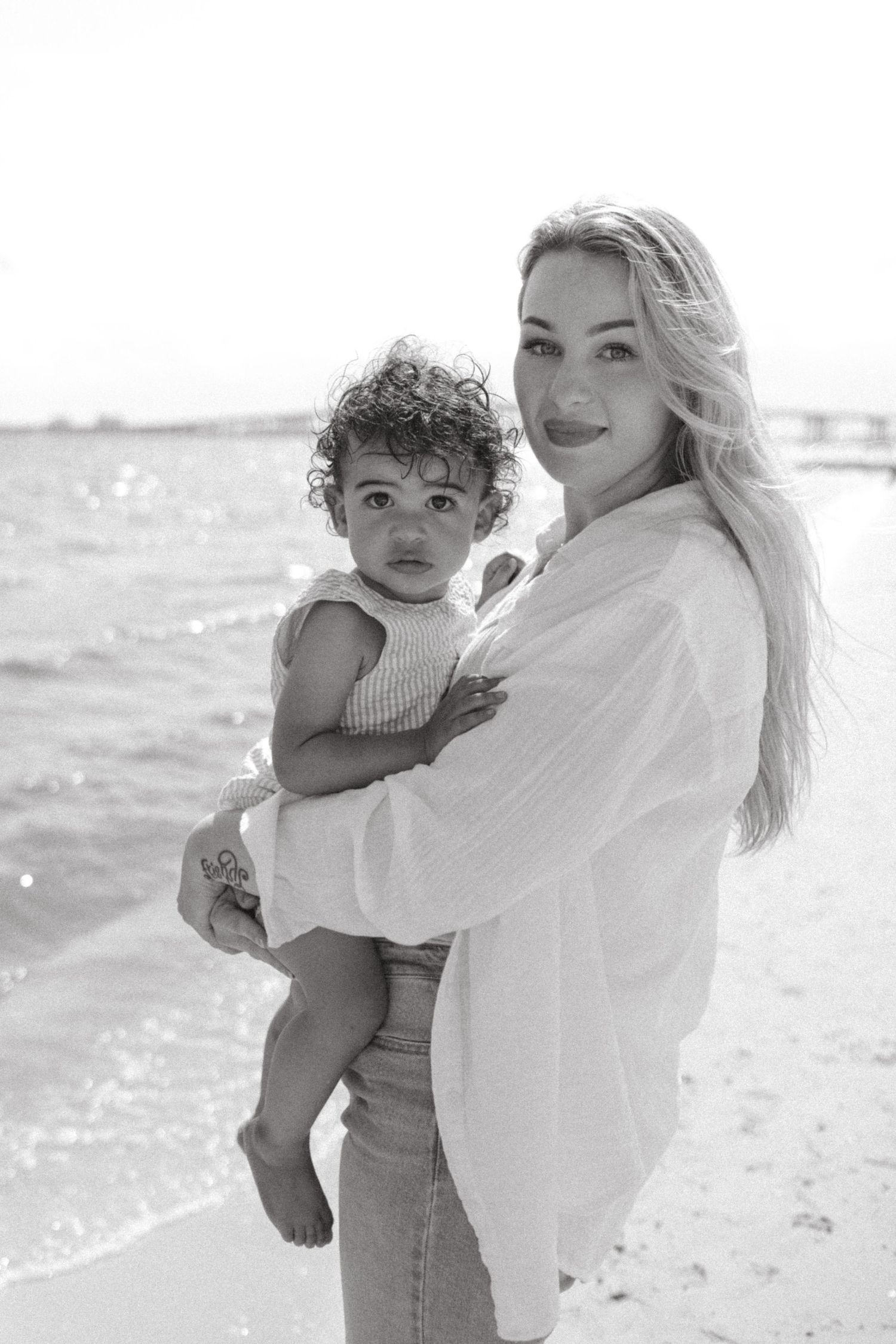Black and white photo of a parent and child embracing on a sunny beach with waves in the background.