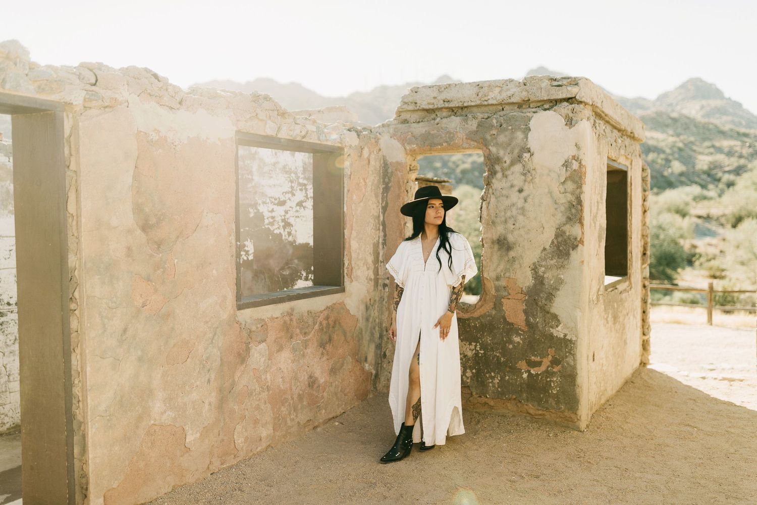 Figure in a white maxi dress and hat stands in stone ruins against desert mountain landscape in warm sunlight.