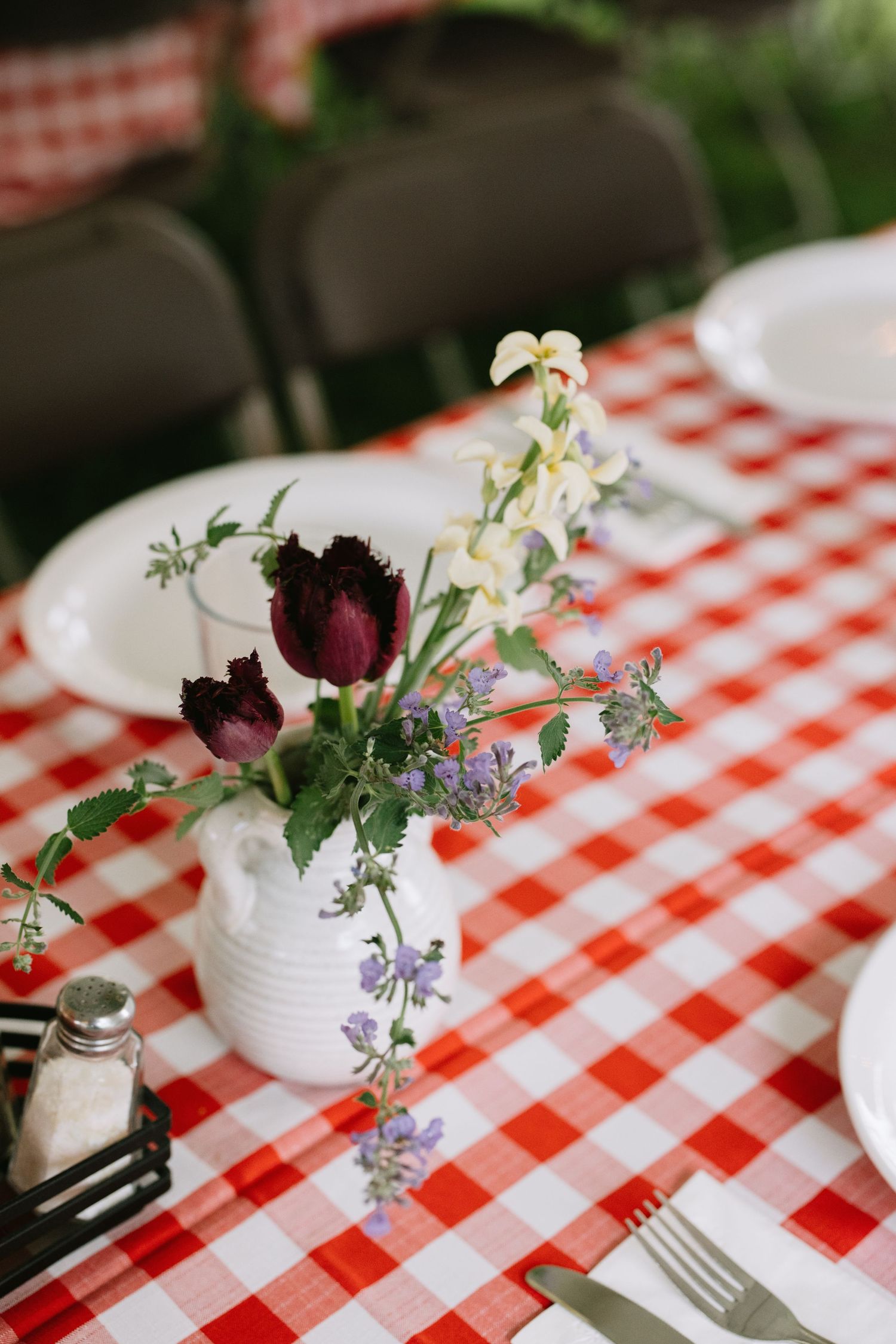 Small white vases with delicate purple and burgundy flowers arranged on a classic red and white checkered tablecloth.