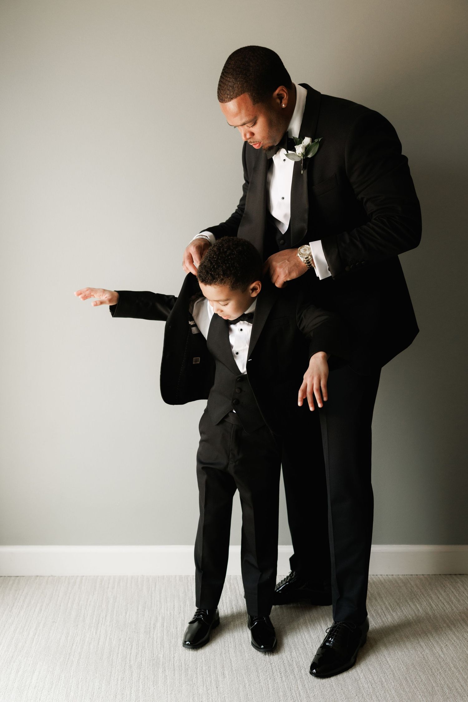 Two people in black suits stand close together in emotional poses against a light-colored wall in a minimal setting.