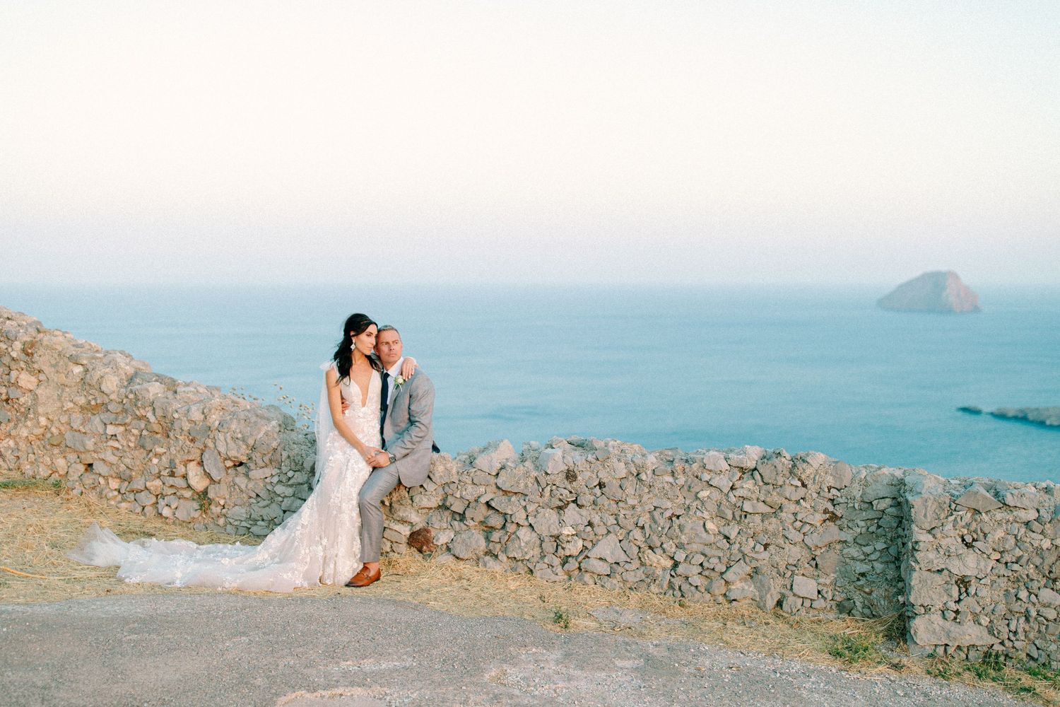 A couple in wedding attire embraces on a rocky coastal cliff overlooking turquoise ocean waters at sunset.