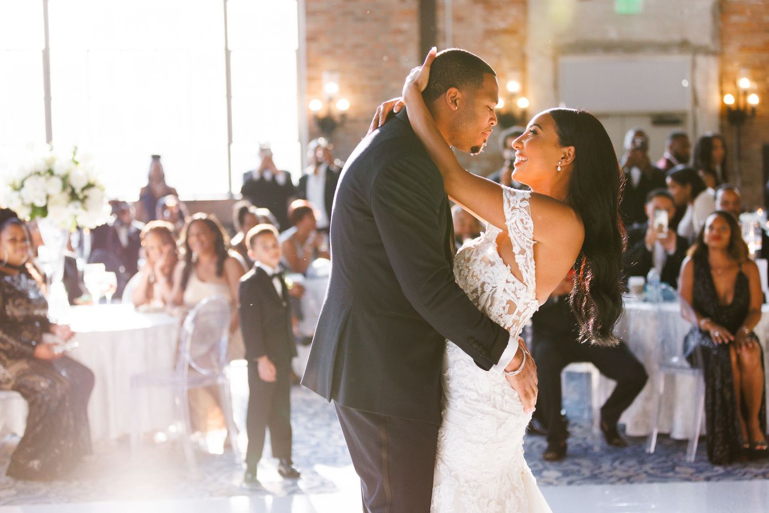 A couple shares their first dance as newlyweds in an elegant ballroom filled with guests and natural light.