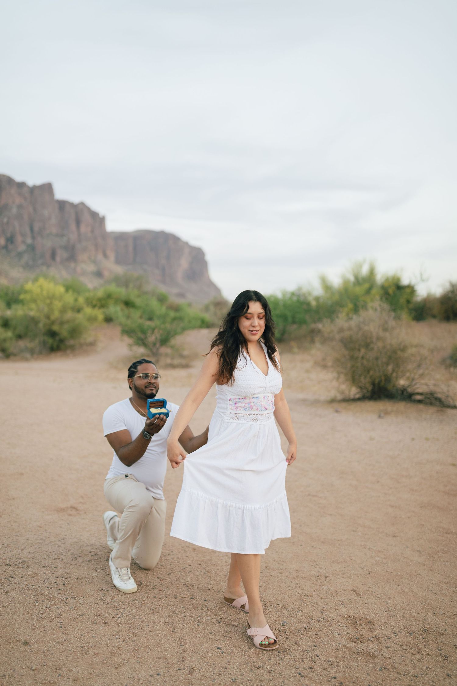Couple engaged in a surprise proposal moment on a desert trail with mountain cliffs in the background.