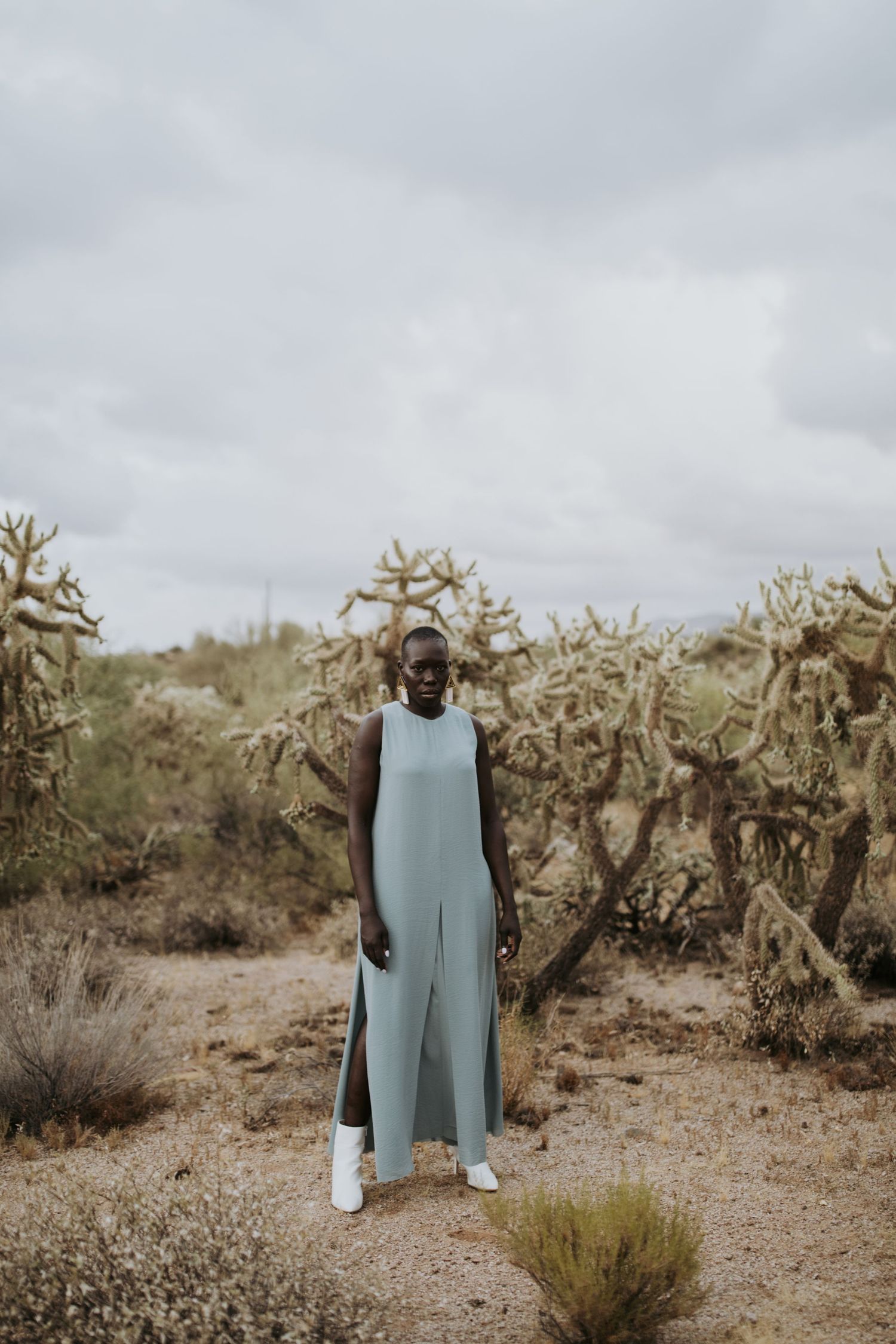 A person wearing a long light blue jumpsuit stands in front of tall desert cactus under an overcast sky.
