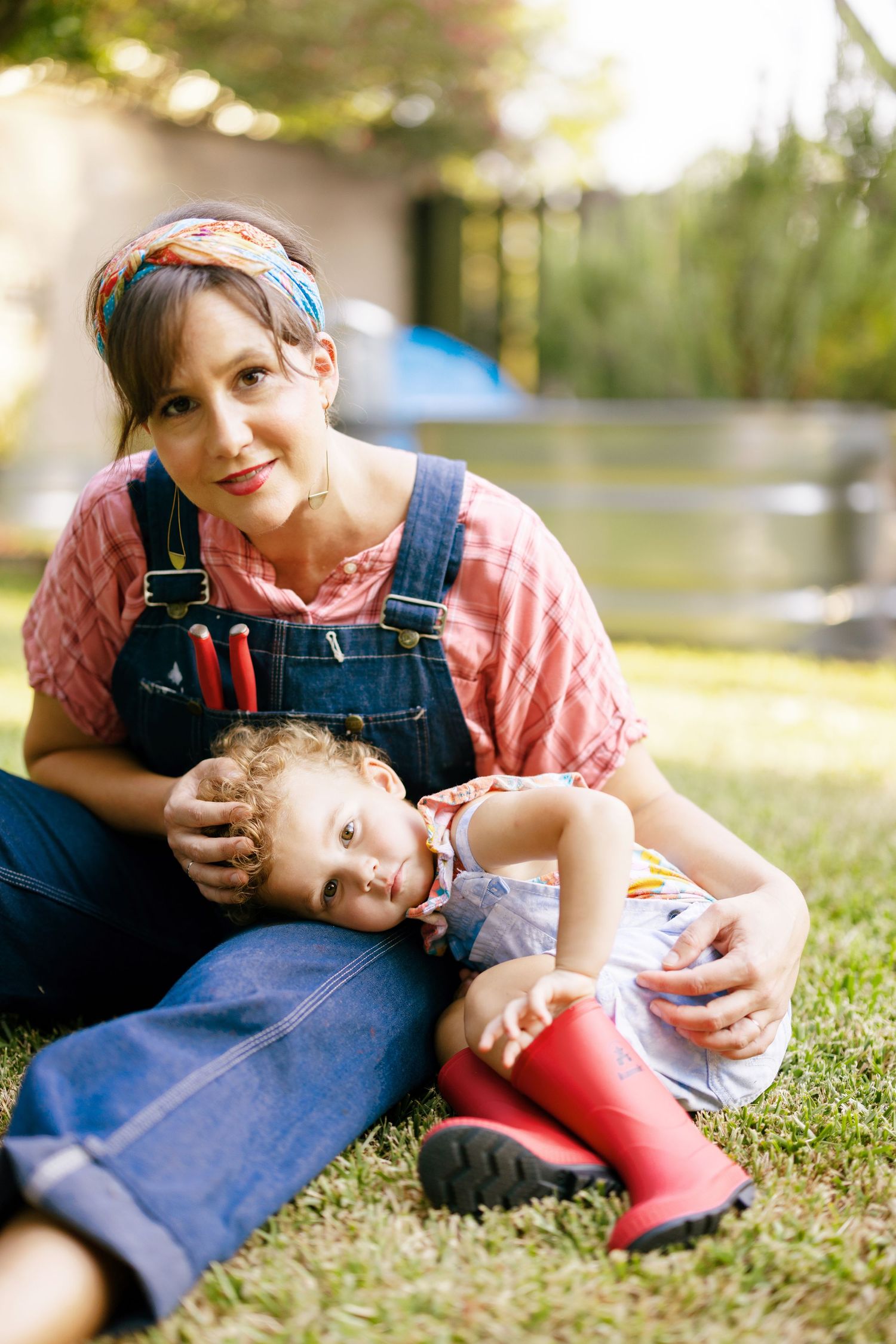Parent and child lie together on grass in a playful candid moment wearing overalls and pink shirt on sunny day.