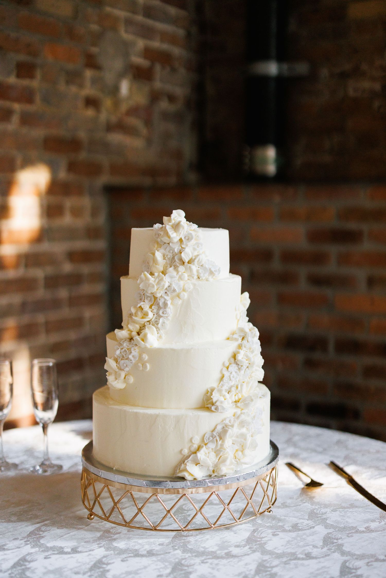 Elegant white wedding cake with delicate floral details sits atop a gold geometric stand against a rustic brick wall backdrop.