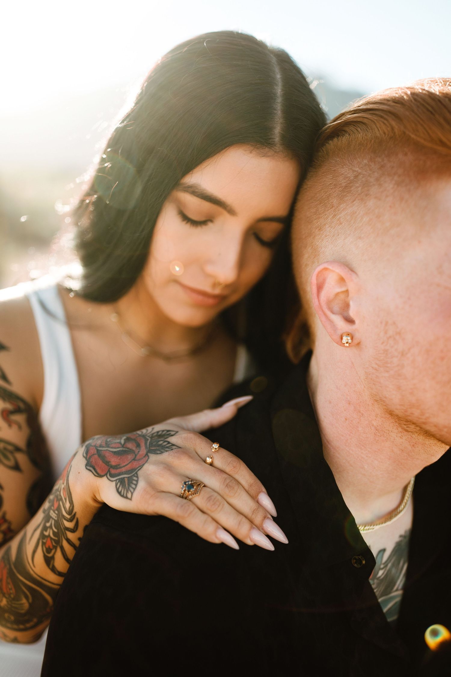 Intimate couple embraces in sunlight with visible tattoos and jewelry creating romantic atmosphere.