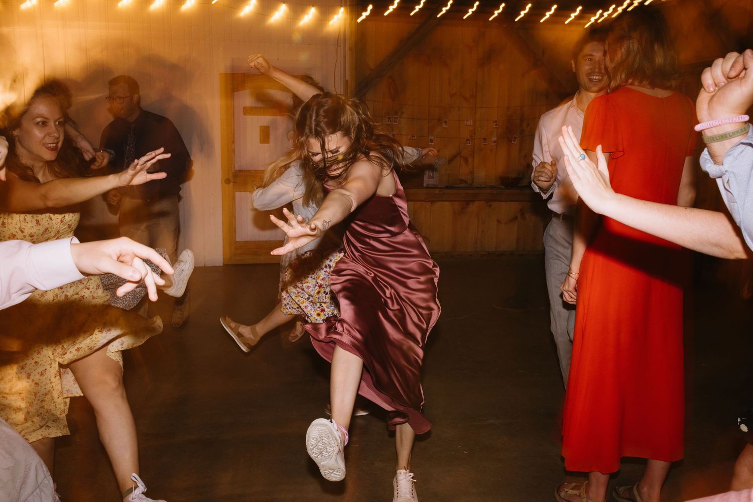 People dance energetically at a party, with a woman in a burgundy dress showing dynamic movement on the dance floor.