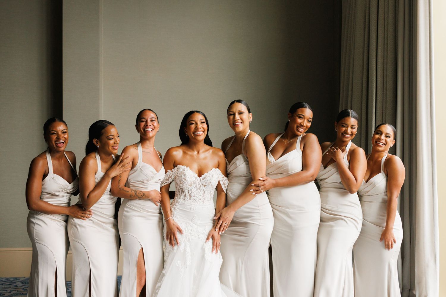 Group of women in matching white dresses gather and laugh together while getting ready for a wedding celebration.