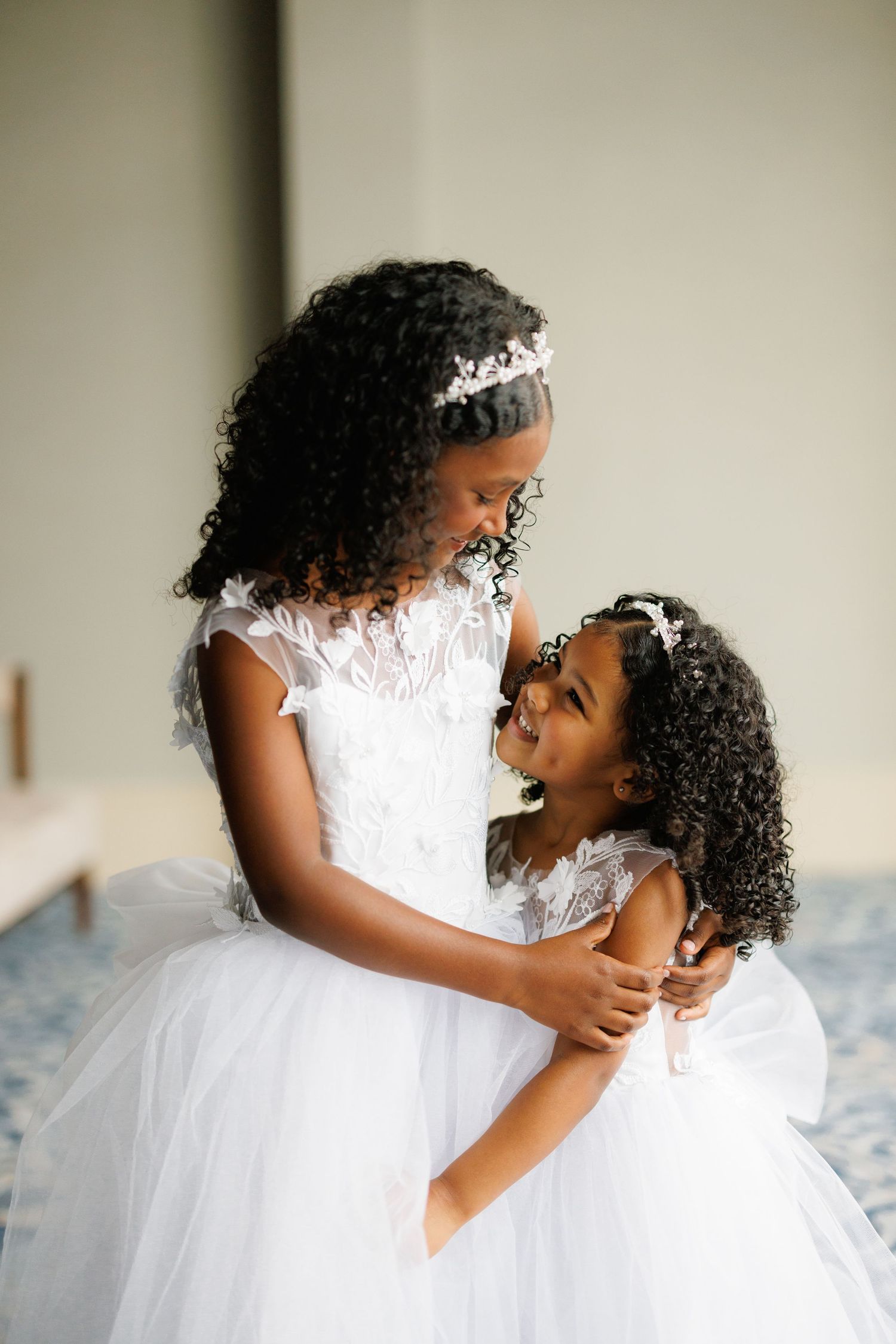 Two children in white tulle dresses and tiaras share a joyful moment together at a formal event.