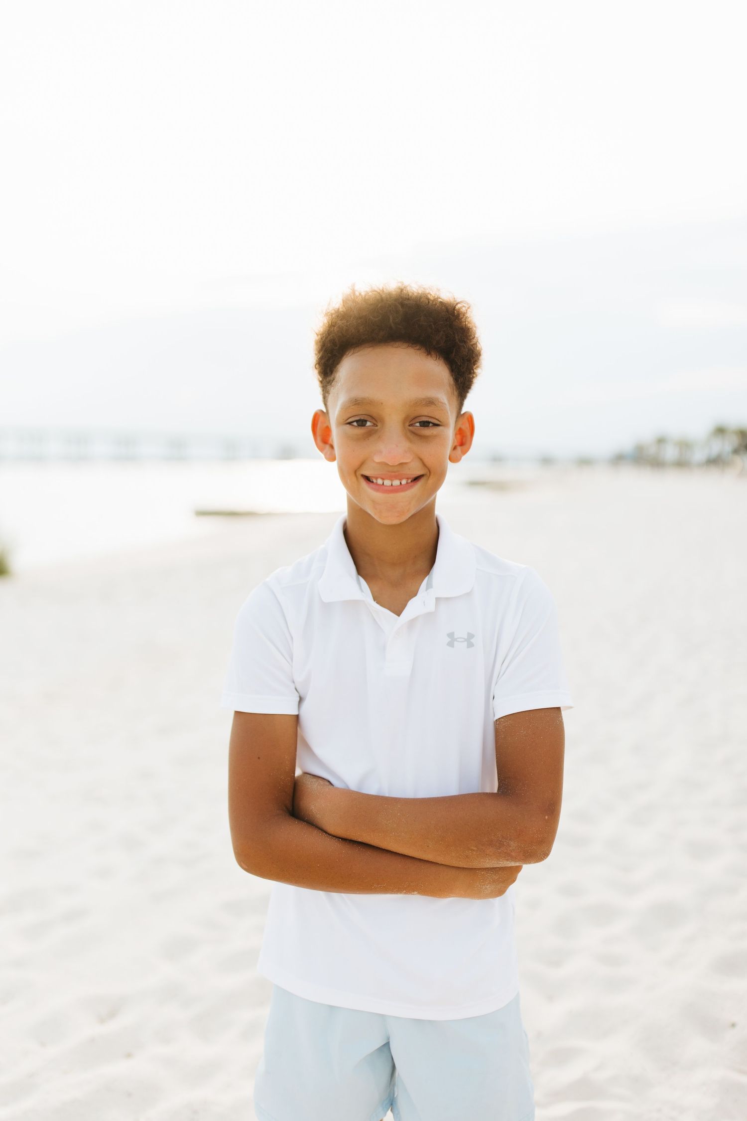 Young person in white polo shirt smiles confidently while standing on beach with ocean view behind.