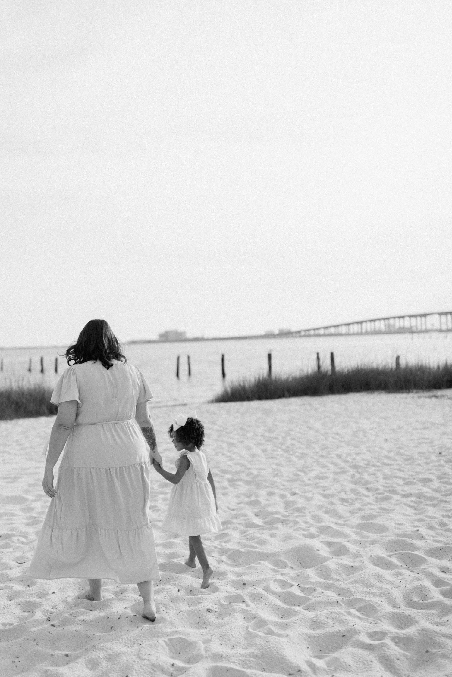 A mother and daughter walk hand in hand on a sandy beach wearing matching white dresses in black and white photography.