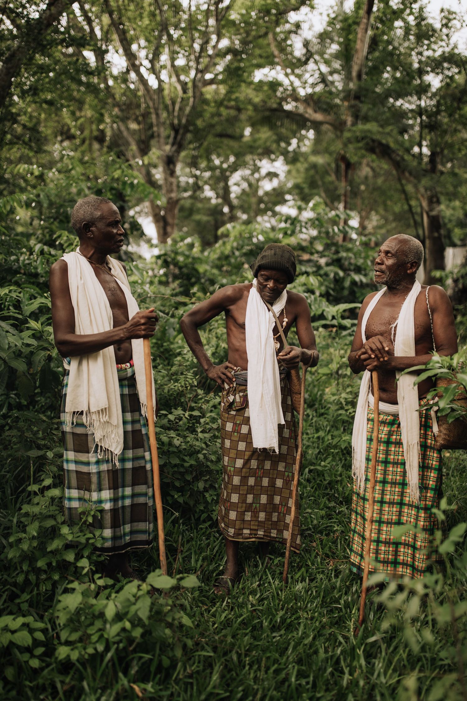 Three people in traditional patterned garments with white scarves stand together outdoors amidst lush greenery.