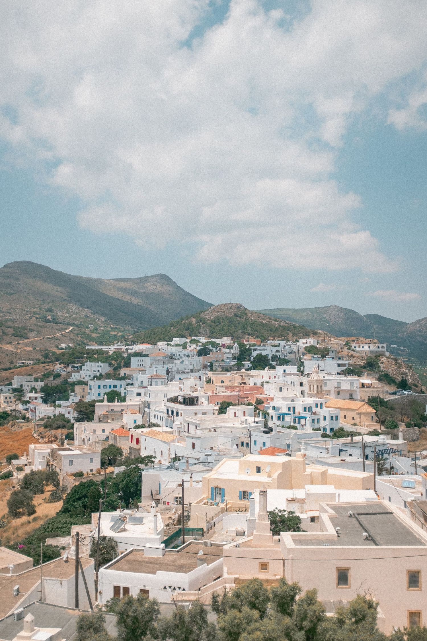 Aerial view of whitewashed buildings nestled in the hills of a Greek island town with mountains in the background.