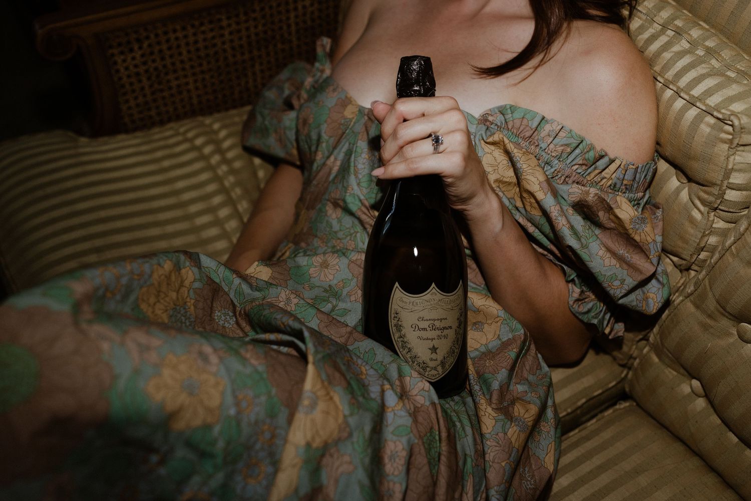 Person in vintage dress holding dark glass bottle while reclining on striped bedding.