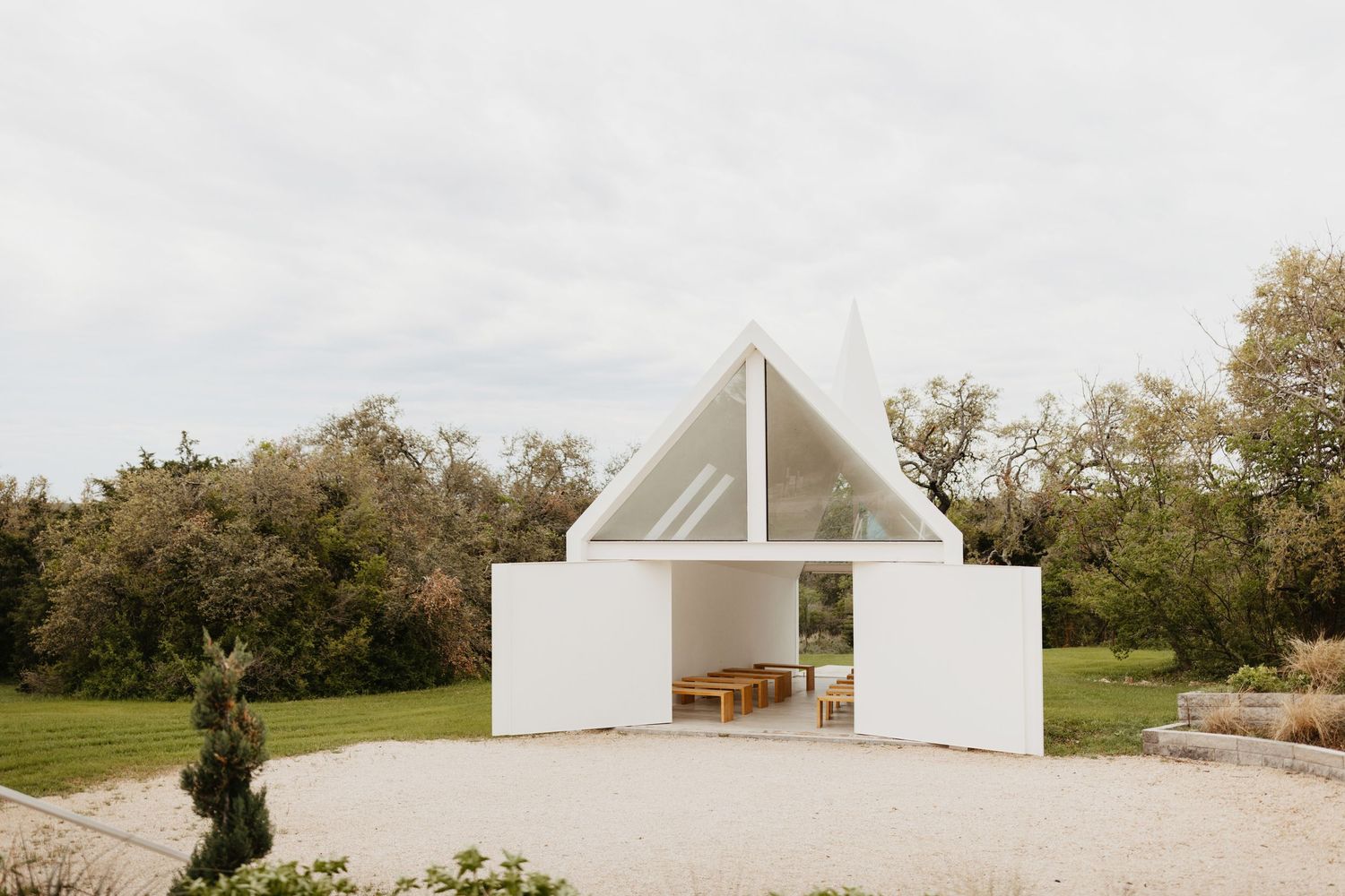 Minimalist white chapel with triangular roof and large doors stands in a serene garden setting.