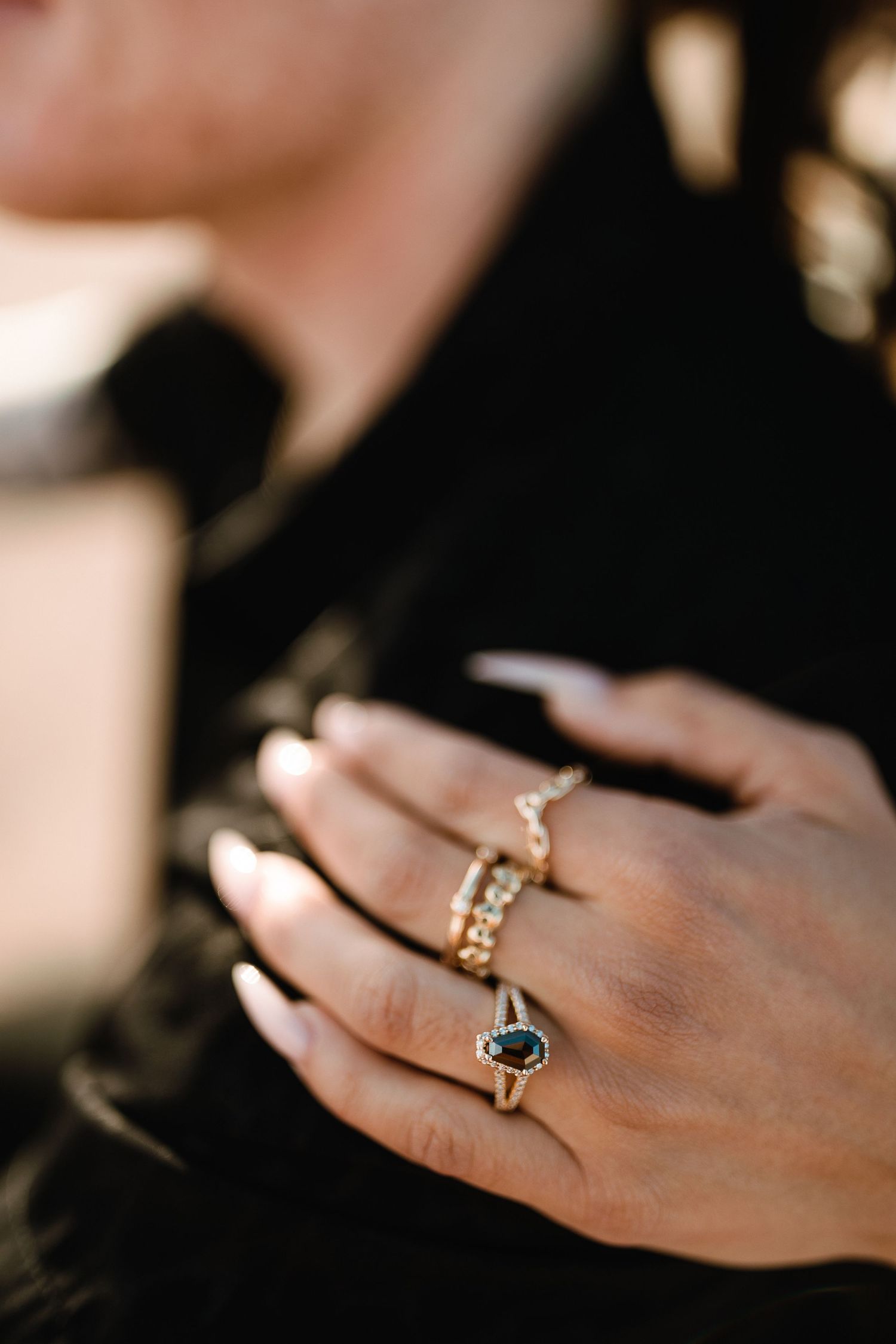 Close up of a hand wearing multiple delicate gold rings with gemstone details against a dark background.