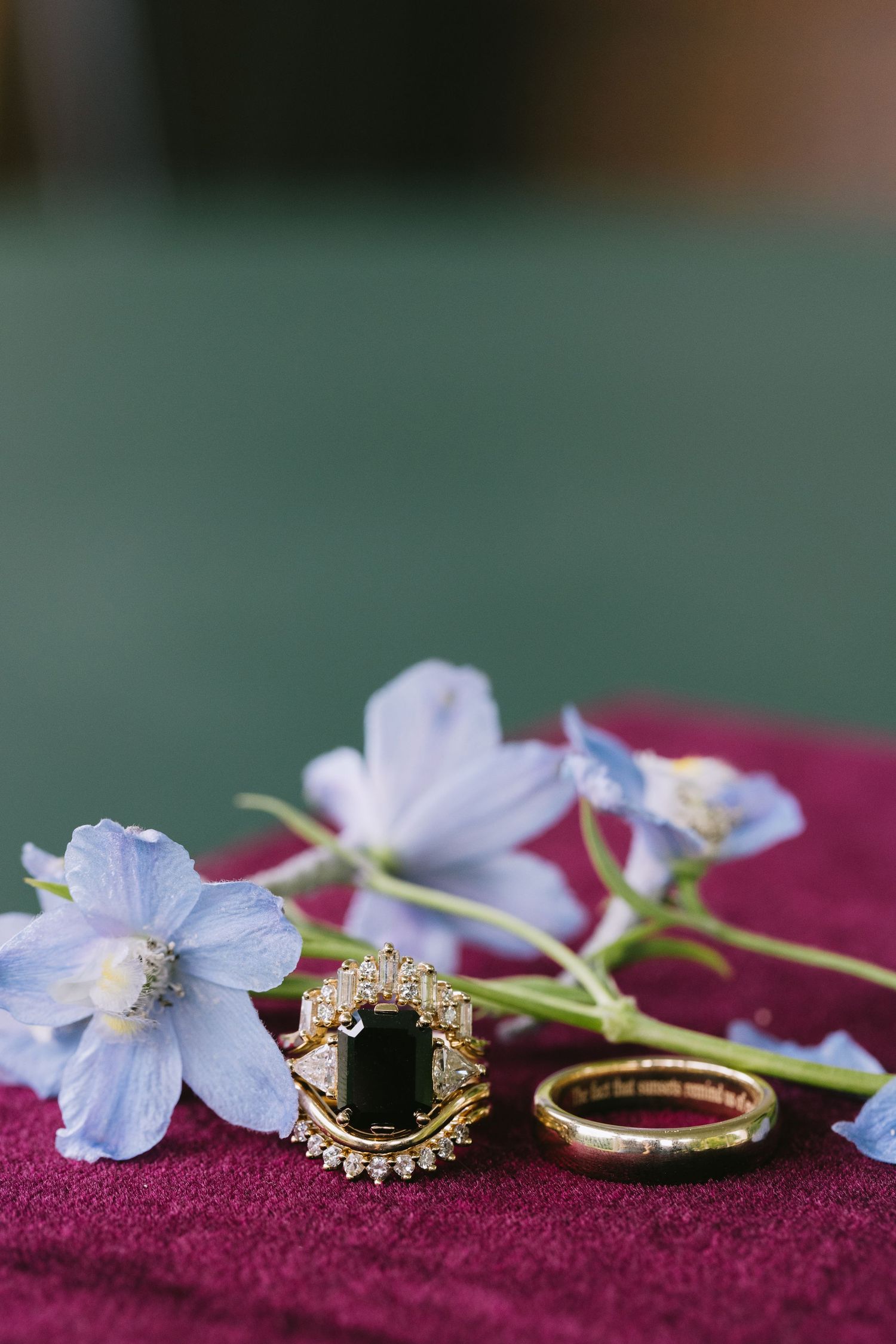 Elegant engagement rings with blue delphinium flowers arranged on burgundy velvet fabric.