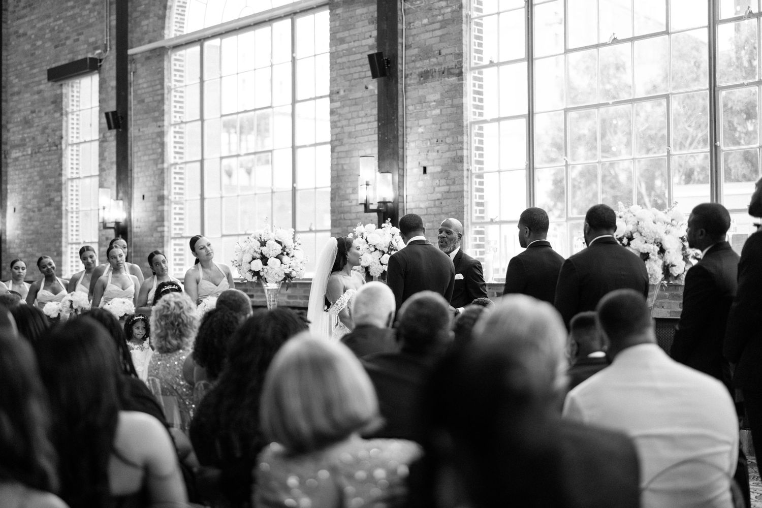 A large crowd of wedding guests gathered in a historic brick venue with tall windows in black and white.