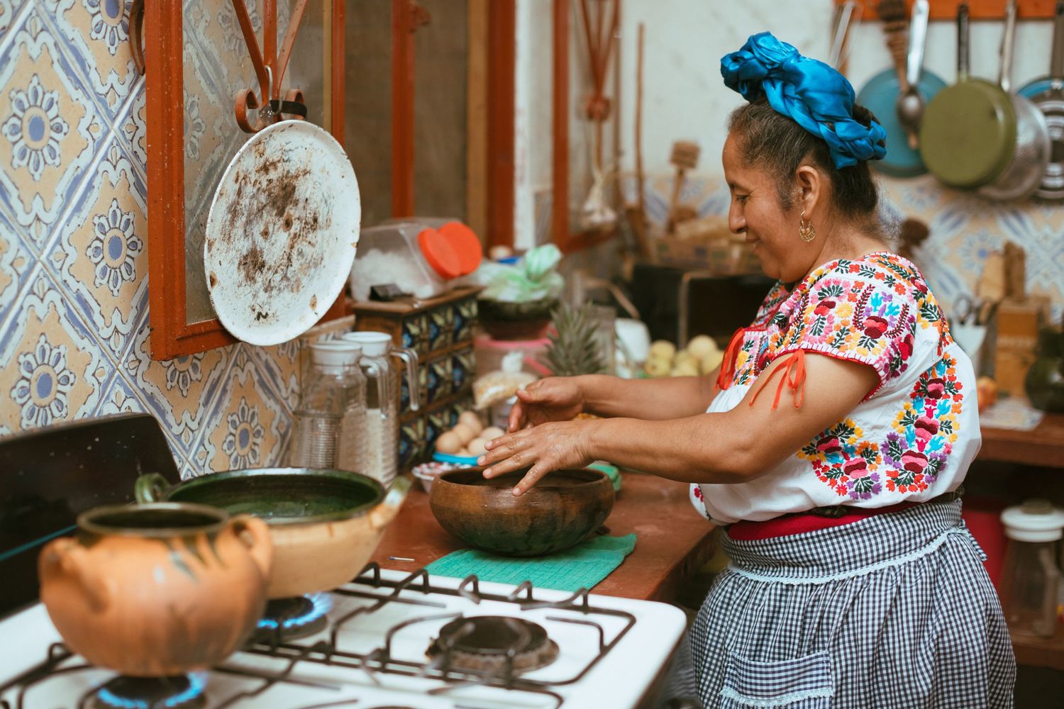 A cook wearing a turquoise headwrap and floral print top prepares food at a traditional stovetop in a rustic kitchen.