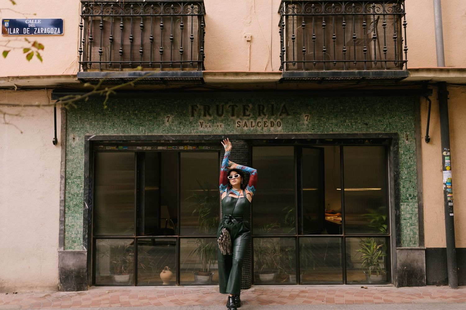 A person in a green jumpsuit stretches to touch a rustic building facade with ornate iron balconies in an urban setting.