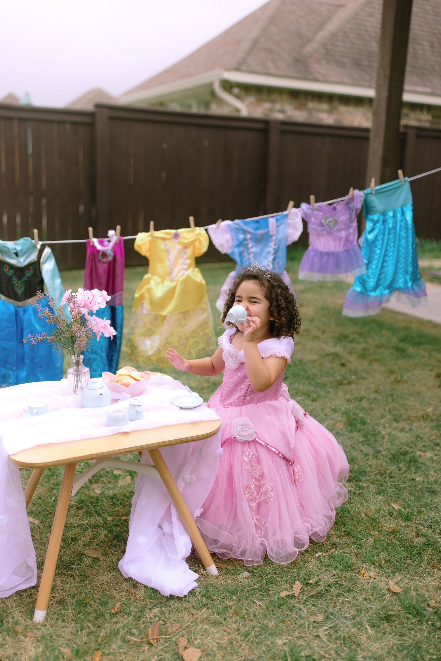 A child in a pink tulle dress has an outdoor tea party under a clothesline of colorful garments hanging to dry.