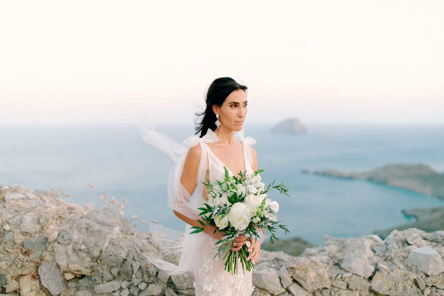 Scenic wedding couple poses on mountain cliffs overlooking a serene coastal bay with white floral bouquet in hand.