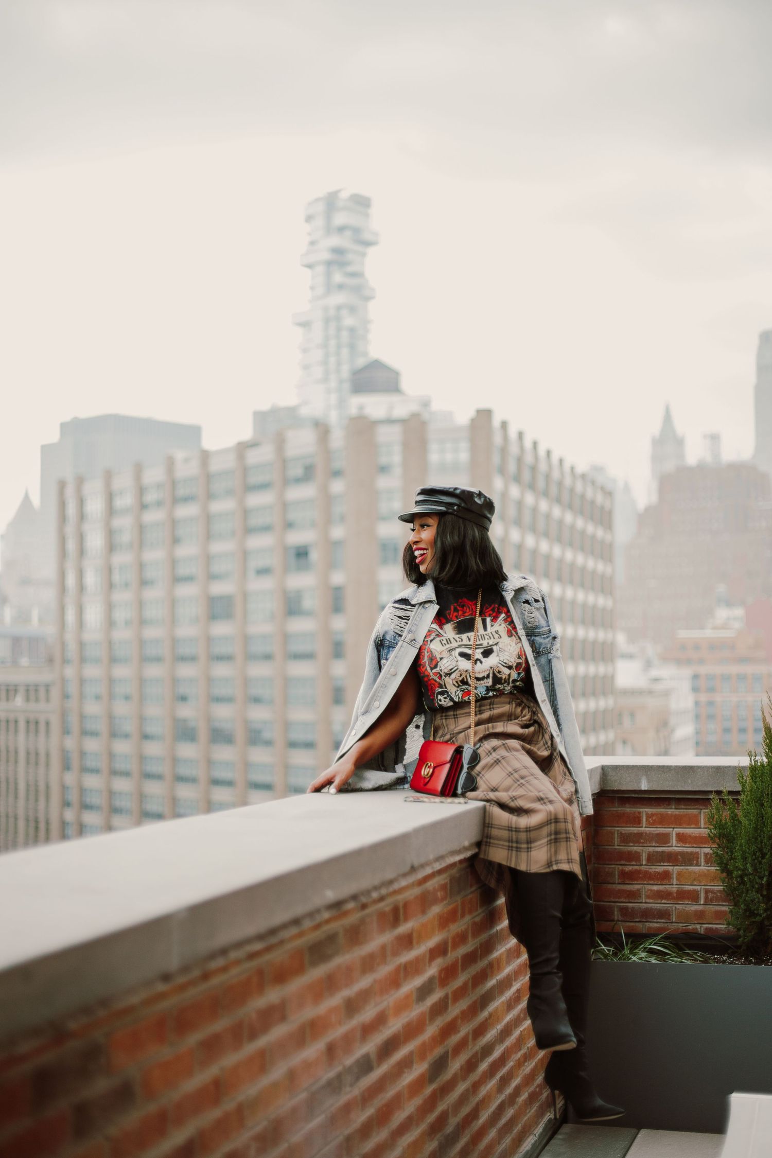 Stylish person in a leather jacket and hat leans against a brick wall overlooking a misty city skyline.