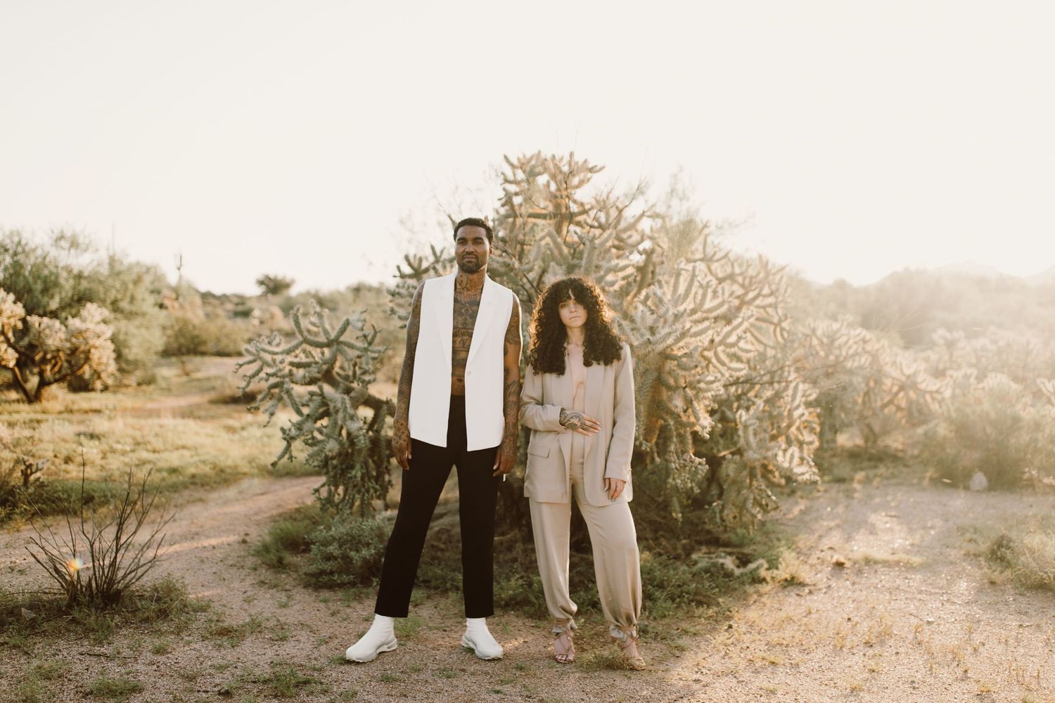 Two people stand together on a desert path surrounded by desert plants and trees at sunset in warm tones.