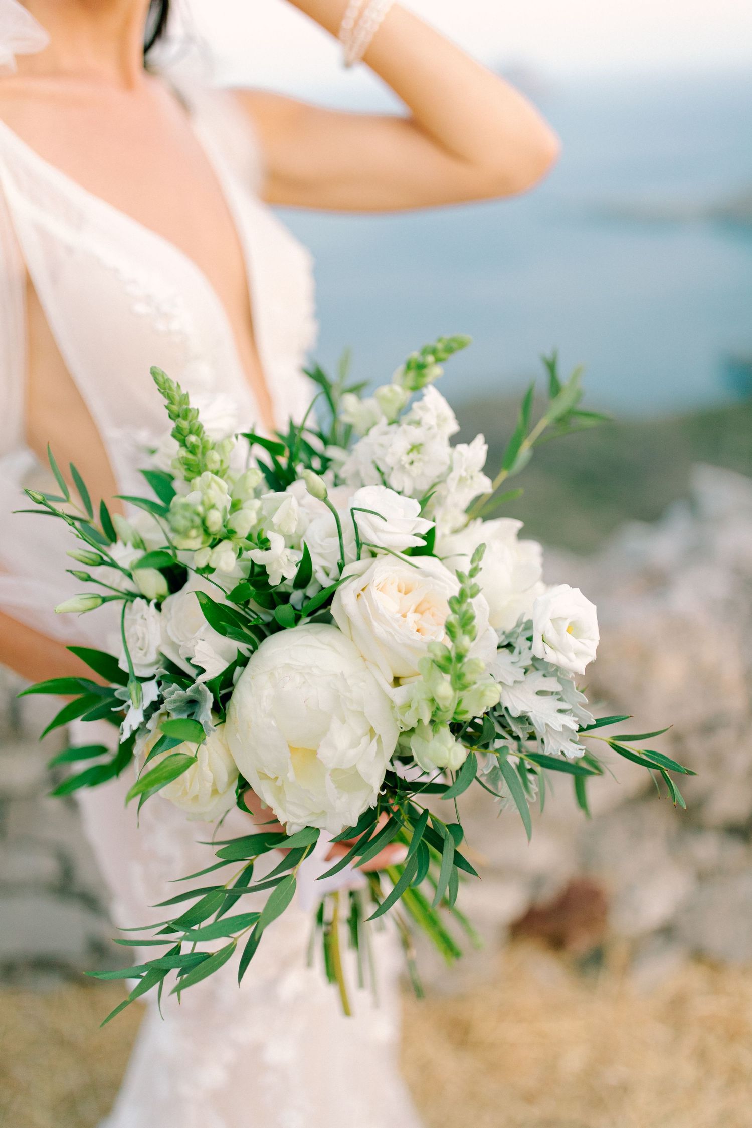 A romantic bridal bouquet with white peonies, roses and fresh greenery held against a soft outdoor backdrop.
