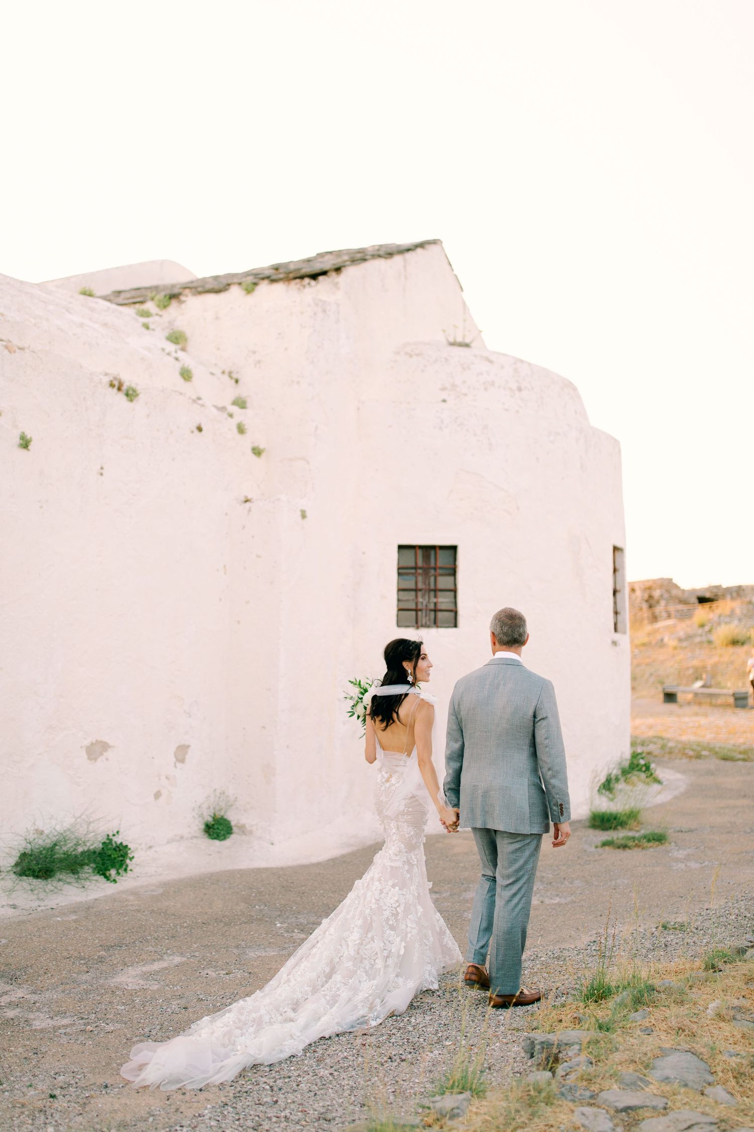 Wedding couple walks hand in hand along rustic white Mediterranean-style building in desert setting at golden hour.