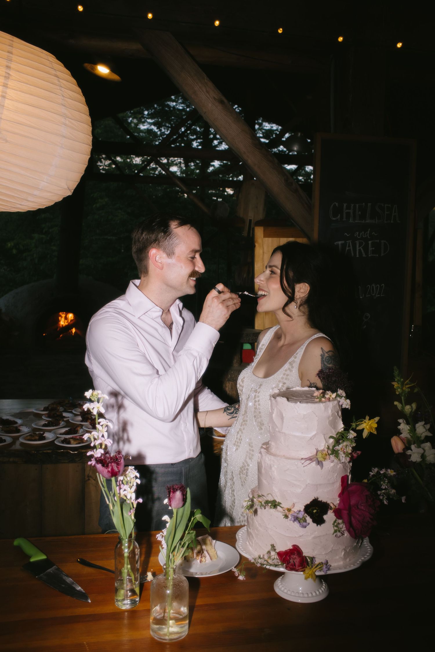 A couple shares a sweet moment feeding each other wedding cake at a rustic reception with paper lanterns and floral decor.