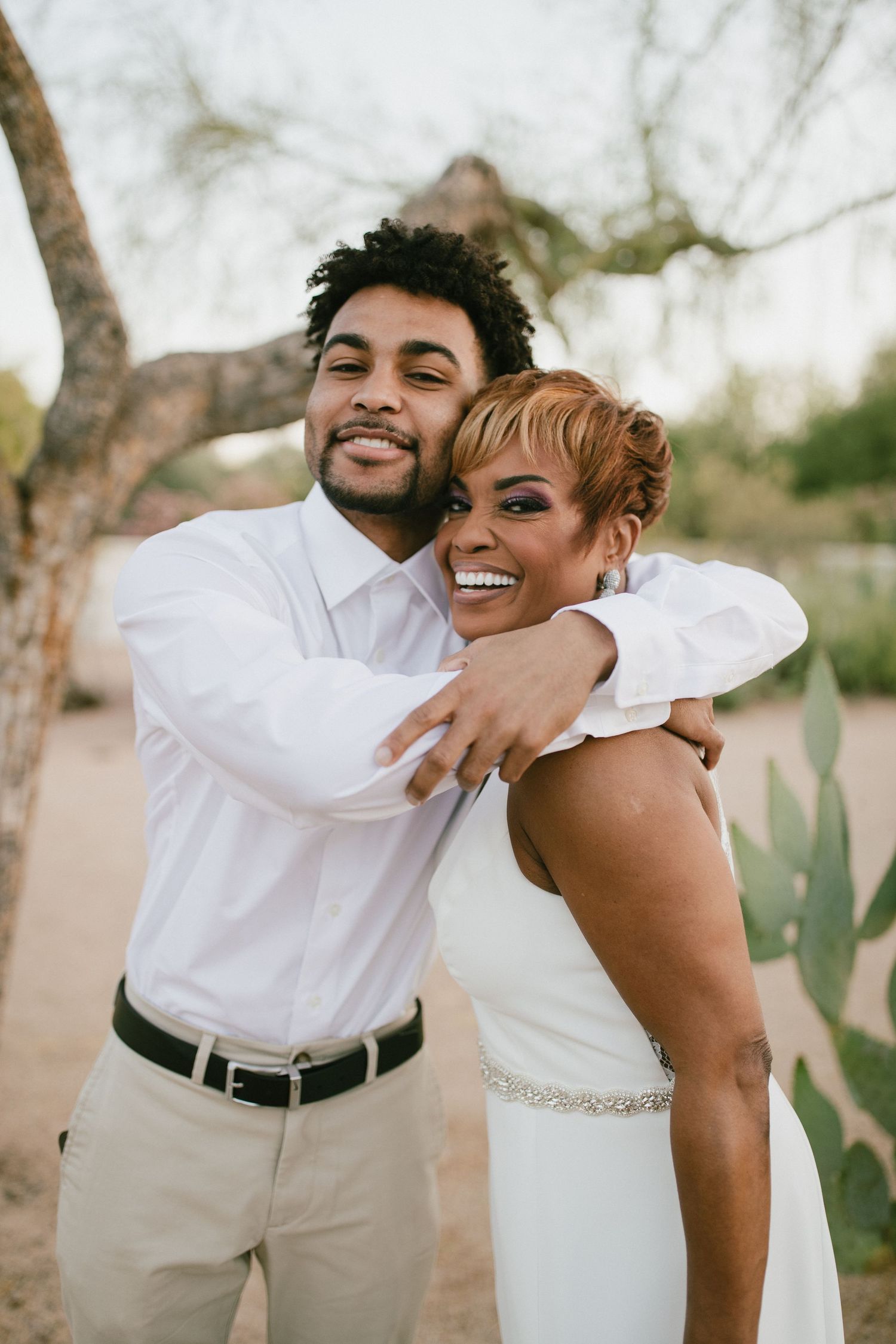 A joyful couple shares a loving embrace outdoors in wedding attire with khaki pants and white clothing.