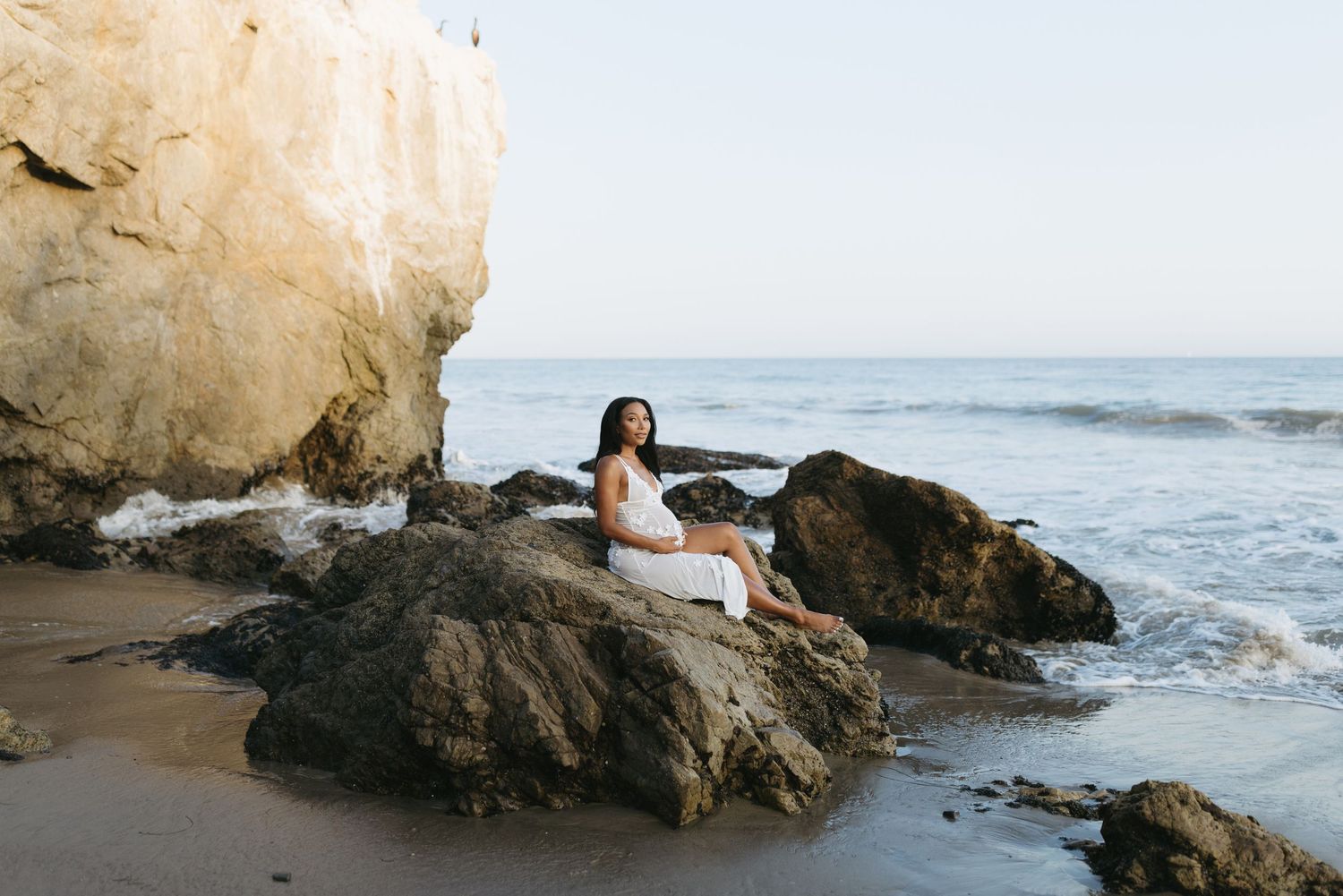 Person relaxing against coastal rocks as waves crash on a scenic beach at sunset.