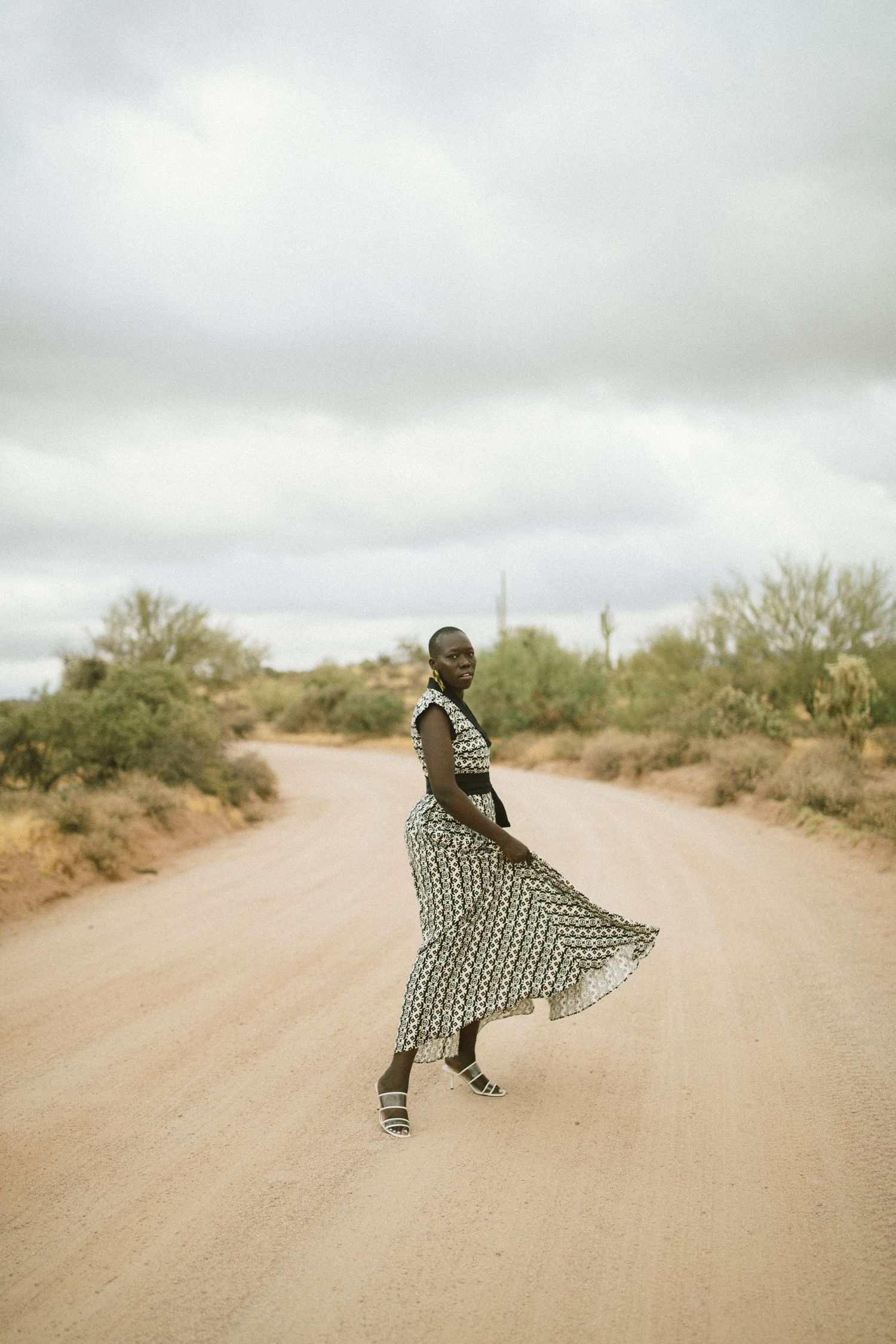 A figure in a flowing patterned dress twirls gracefully on a dusty desert road beneath cloudy skies.