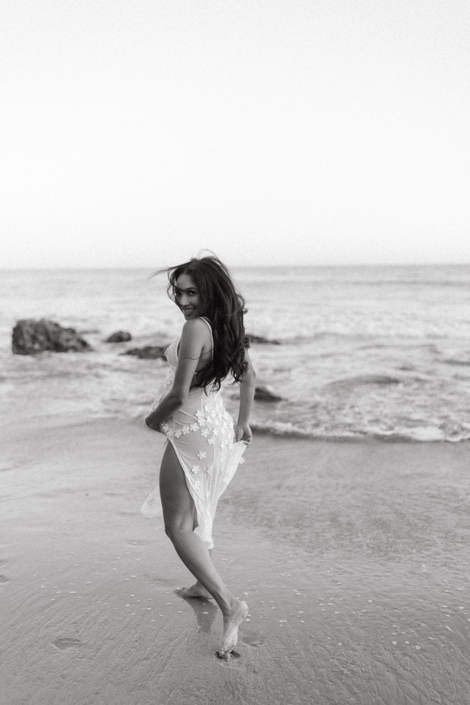 A graceful figure in a flowy white dress dances playfully at the beach shoreline in black and white.