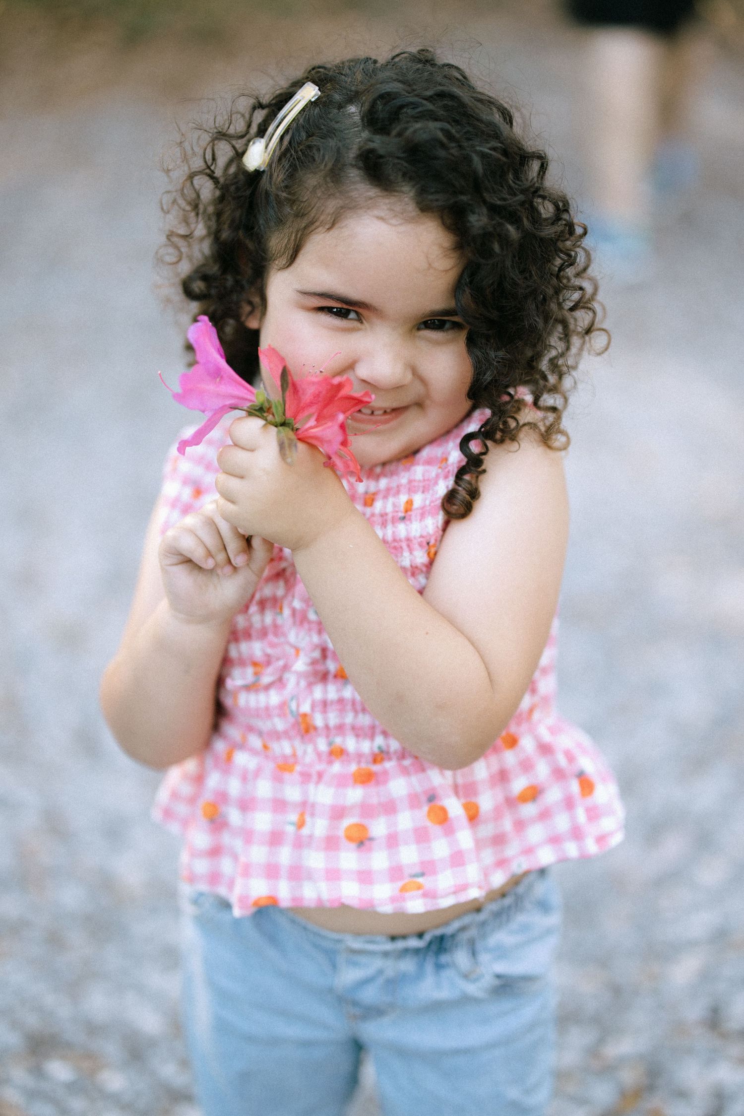 A child with curly hair wearing a pink gingham top and jeans holds a bright pink flower while smiling outdoors.