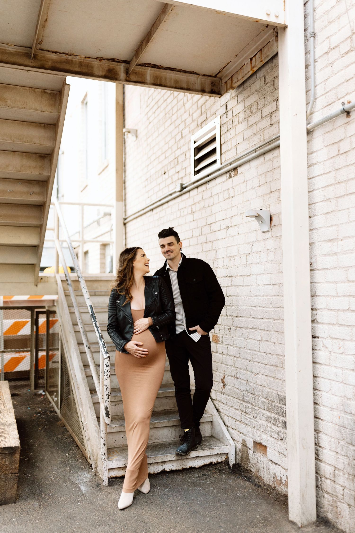 A stylishly dressed couple stands together on white stairs outside an industrial brick building.