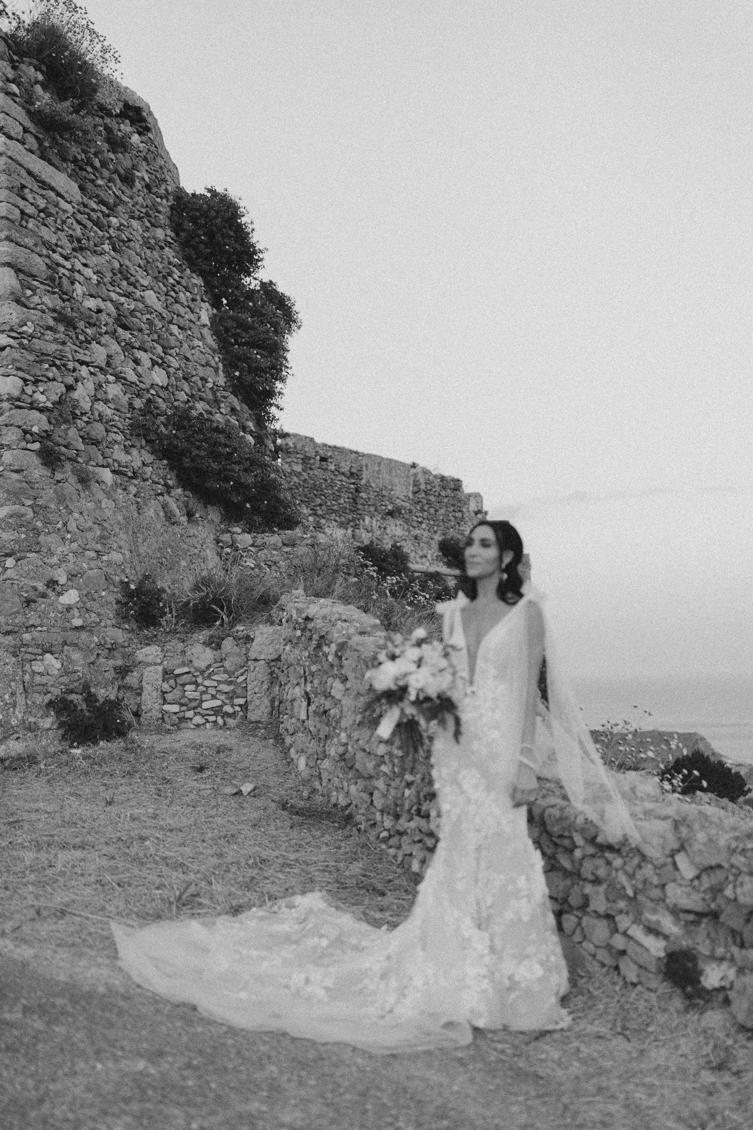 Black and white photo of a bride in lace gown with flowing train standing against ancient stone walls by the sea.