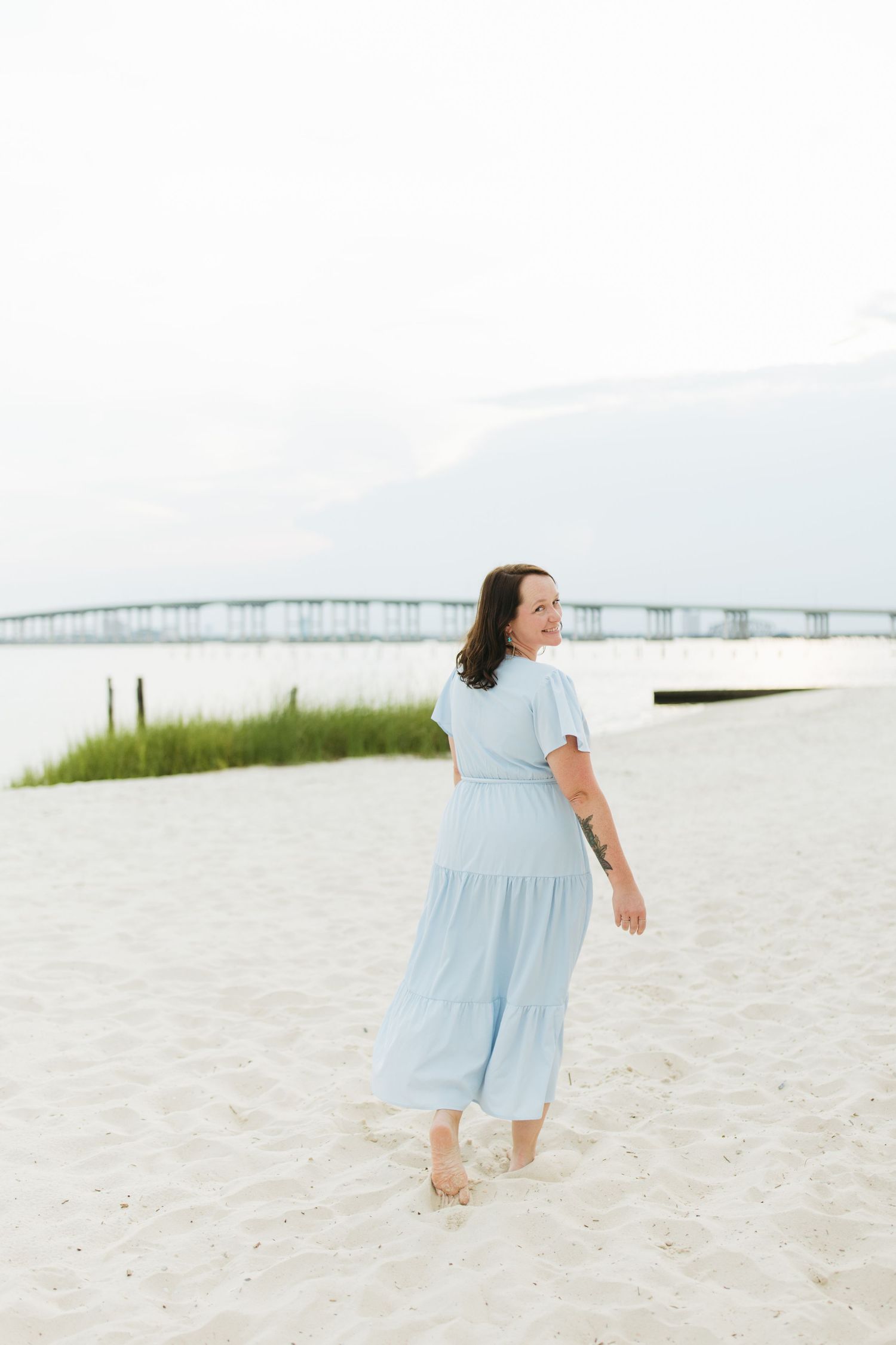 Woman in light blue dress walking barefoot on pristine beach with bridge visible in distance.