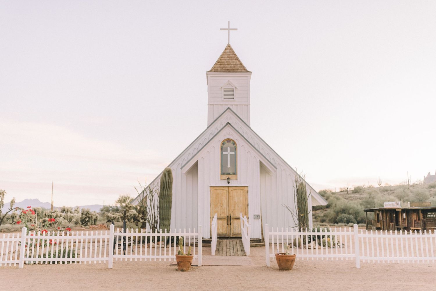 A charming white wooden church with a steeple and picket fence stands against a soft pink sky at sunset.