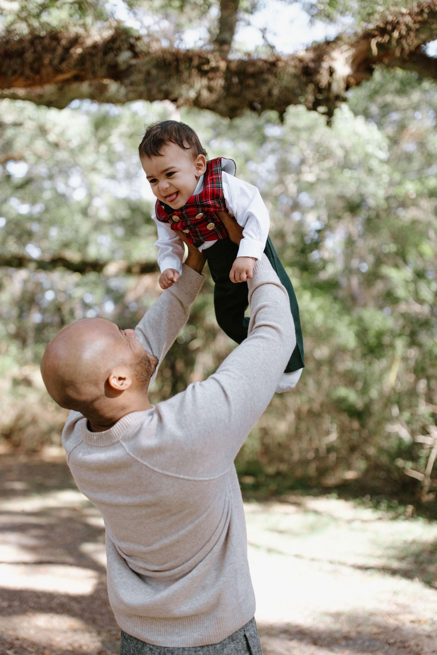 A joyful playtime moment outdoors as an adult lifts a child wearing a red plaid outfit against a natural background.