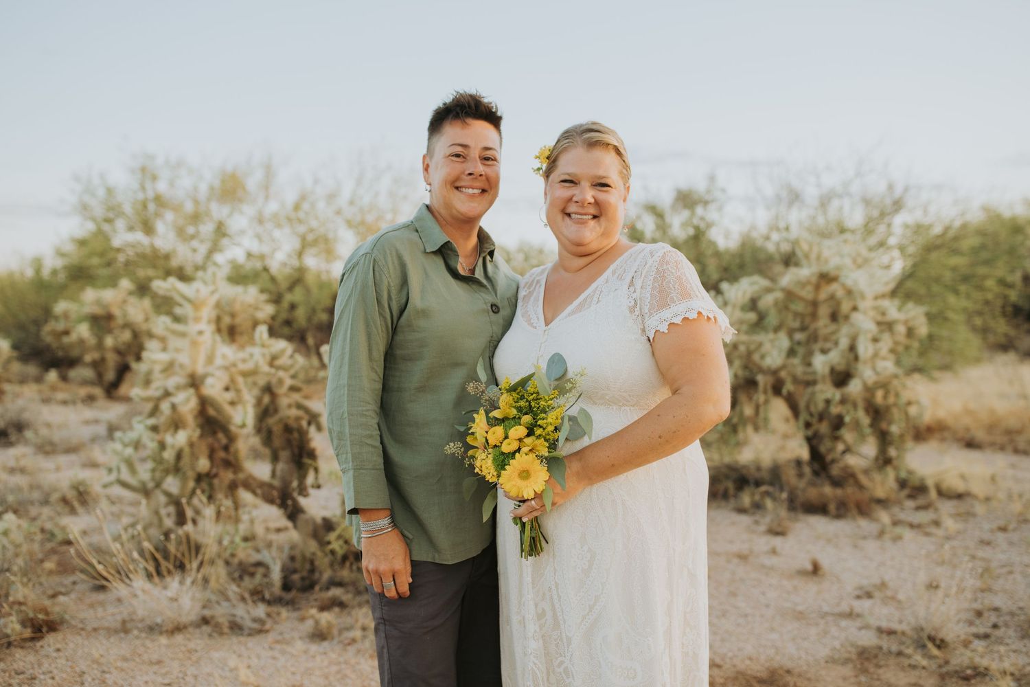 A couple poses together in a desert landscape for wedding photos while holding bright yellow sunflowers.