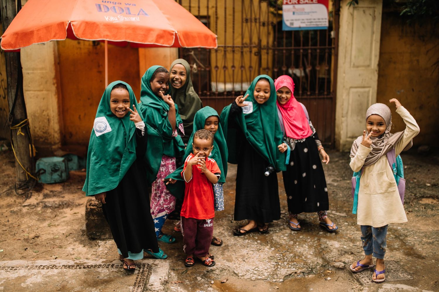 Students in green uniforms and colorful head coverings gather outside under orange umbrellas in the rain.